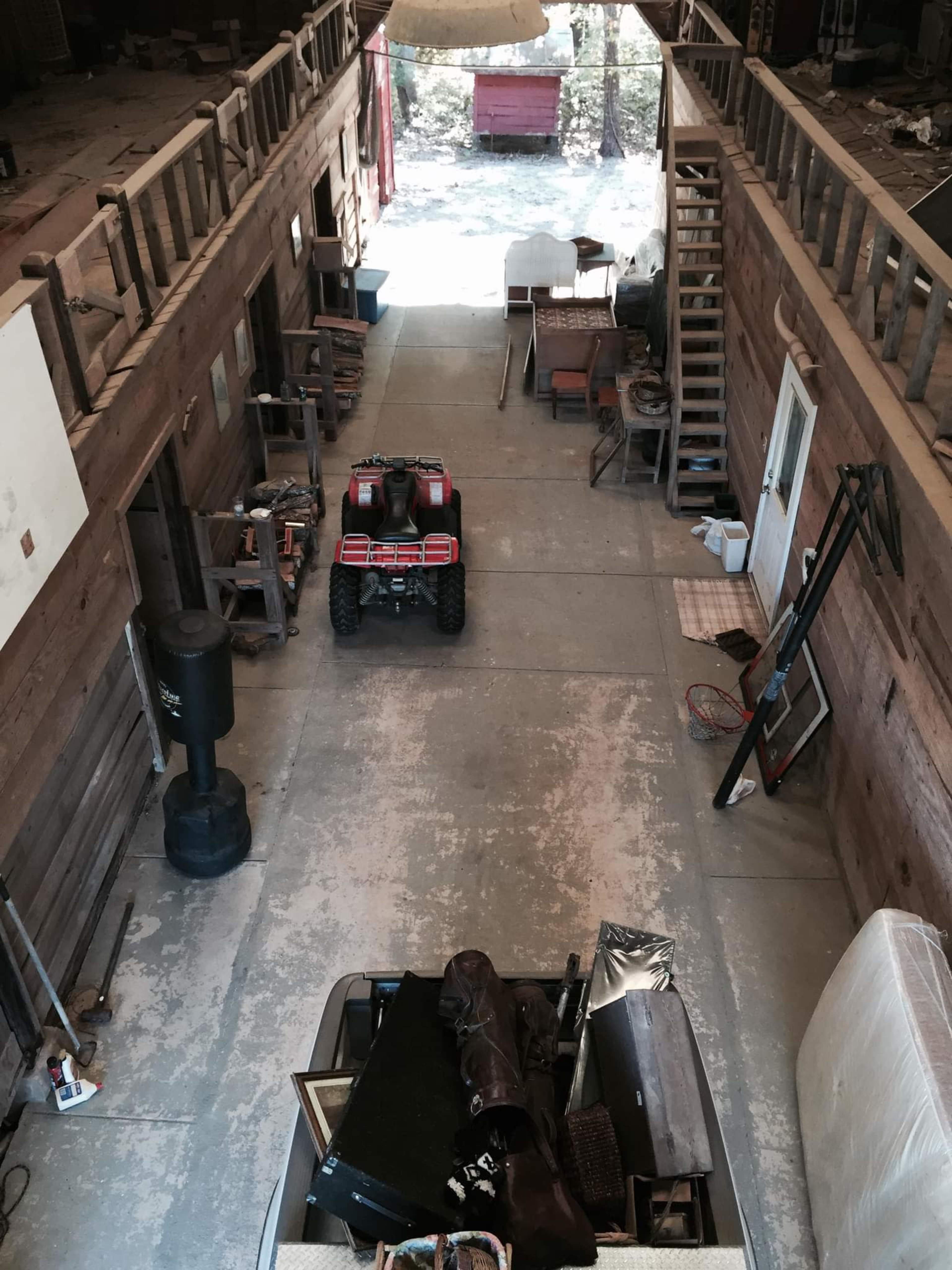 A spacious, unfinished interior of a barn-like structure with an ATV parked in the center, surrounded by various items and wooden beams overhead.