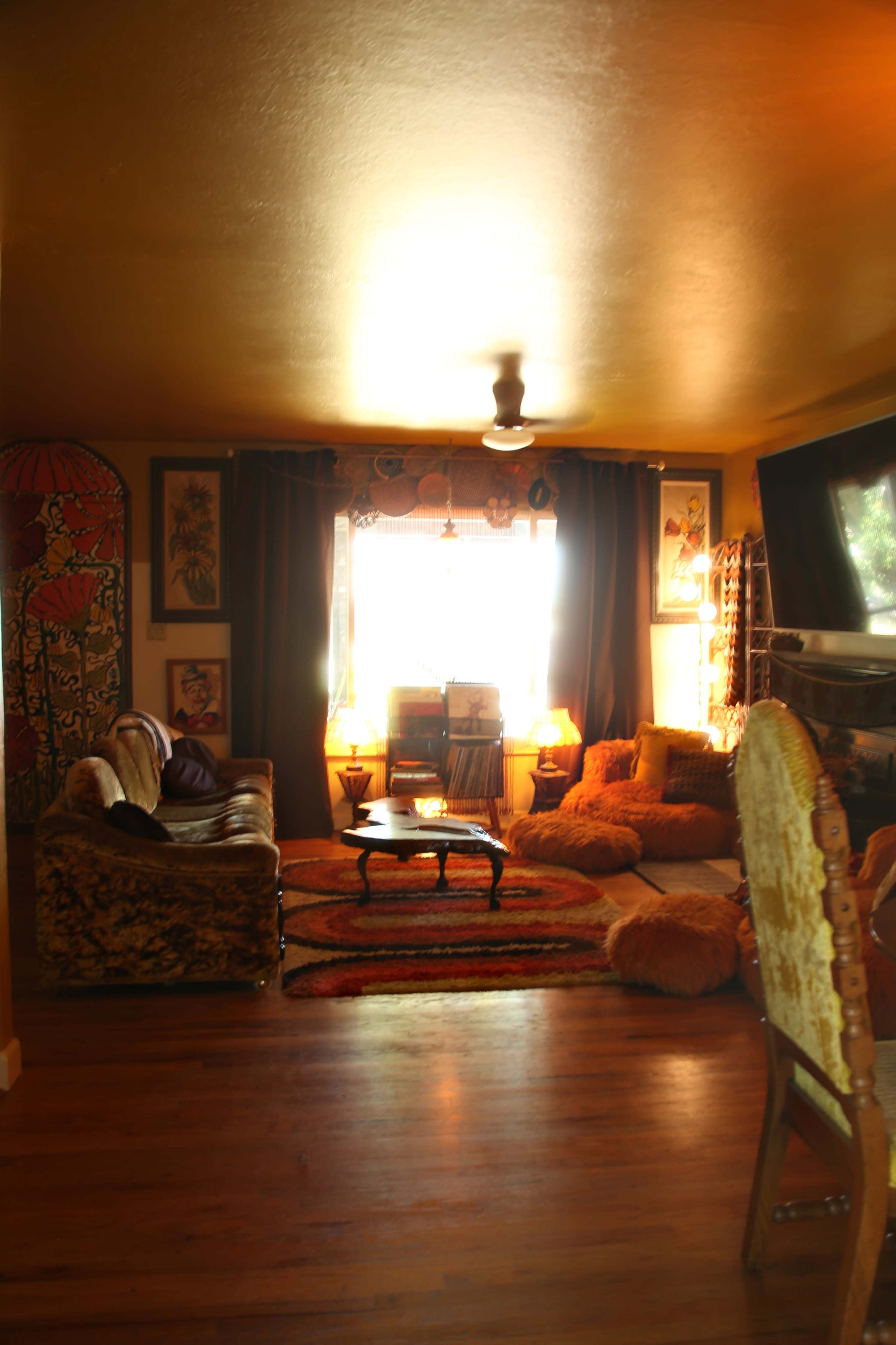 A cozy living room features a patterned couch, a coffee table, floor cushions, and large windows illuminated by natural light.