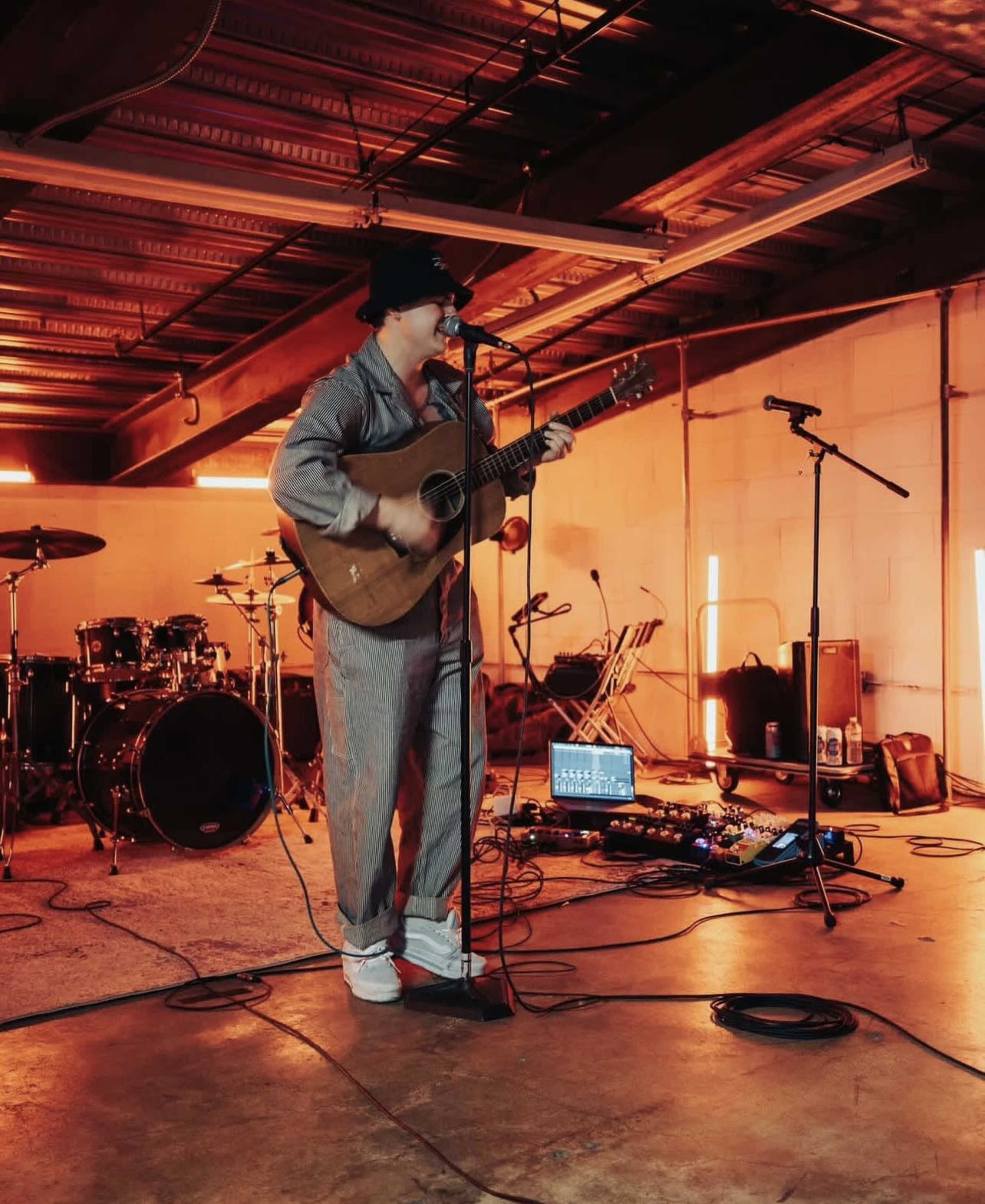A musician in a jumpsuit plays an acoustic guitar on stage in a dimly lit room with musical equipment set up in the background.