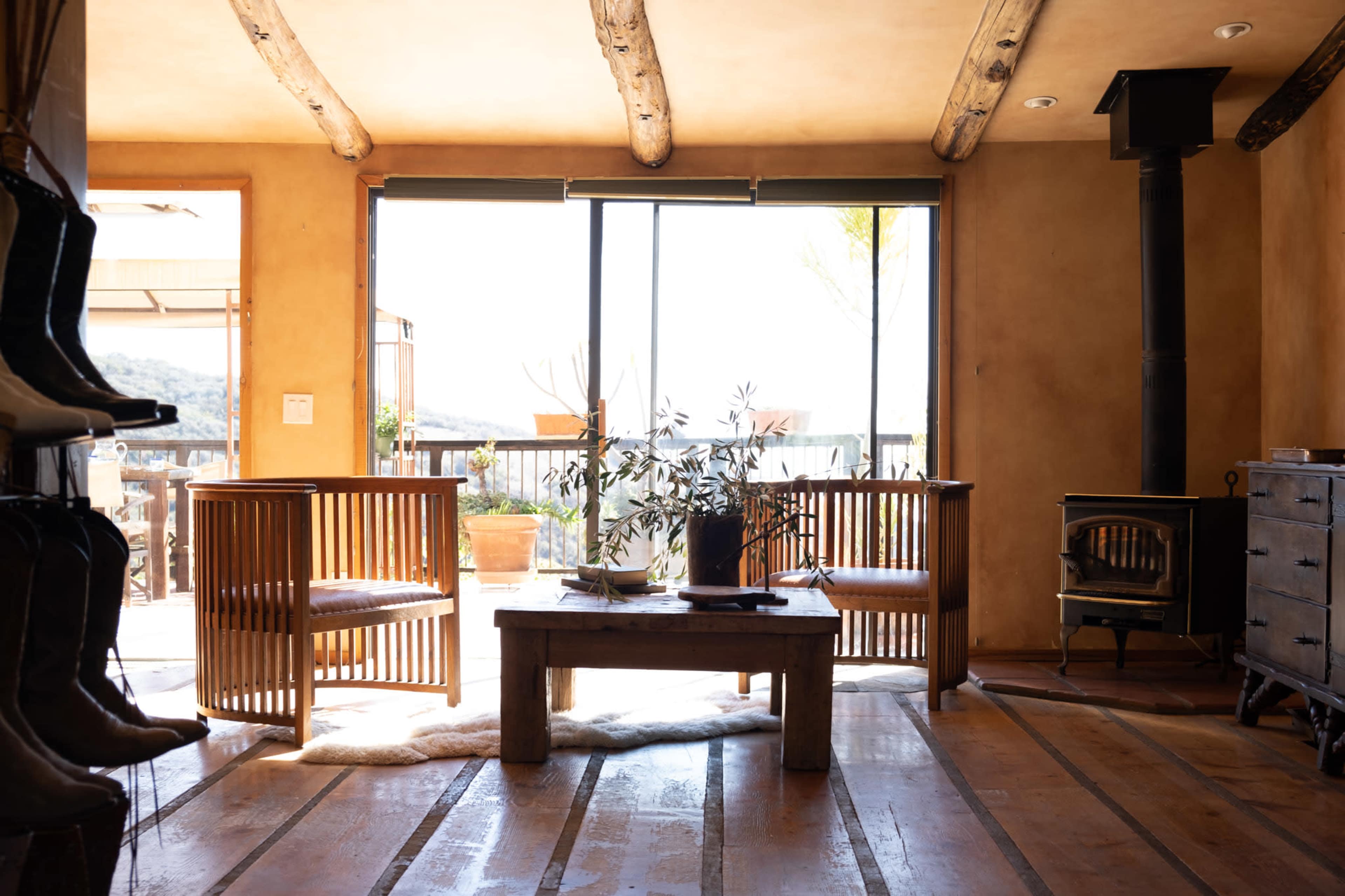 A cozy living room features two wooden chairs facing a table, with large windows providing a view of the outdoors and a wood stove in the corner.