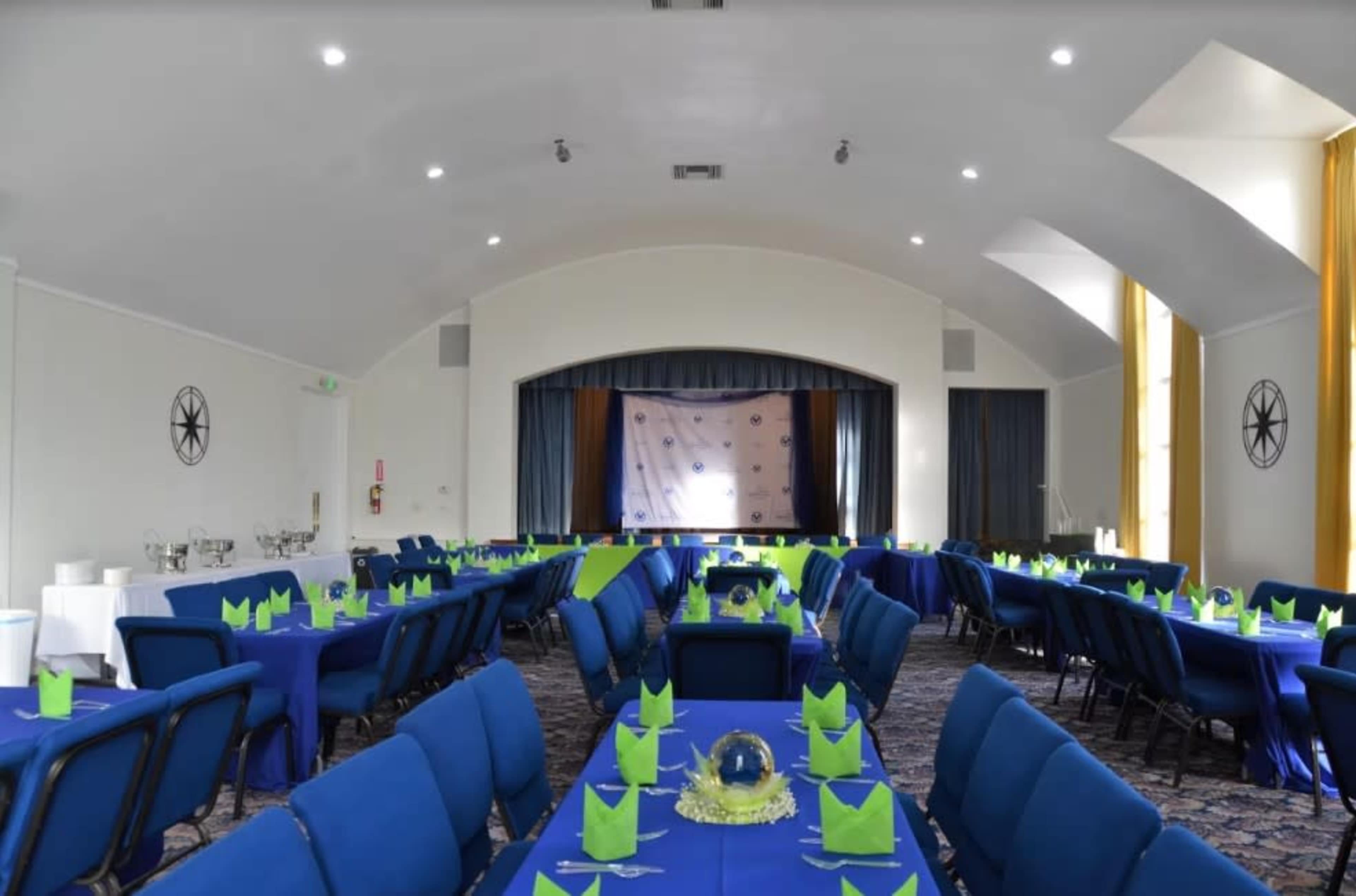 The image shows a banquet hall set up with multiple tables covered in blue cloth, each adorned with green place settings and a decorative centerpiece.