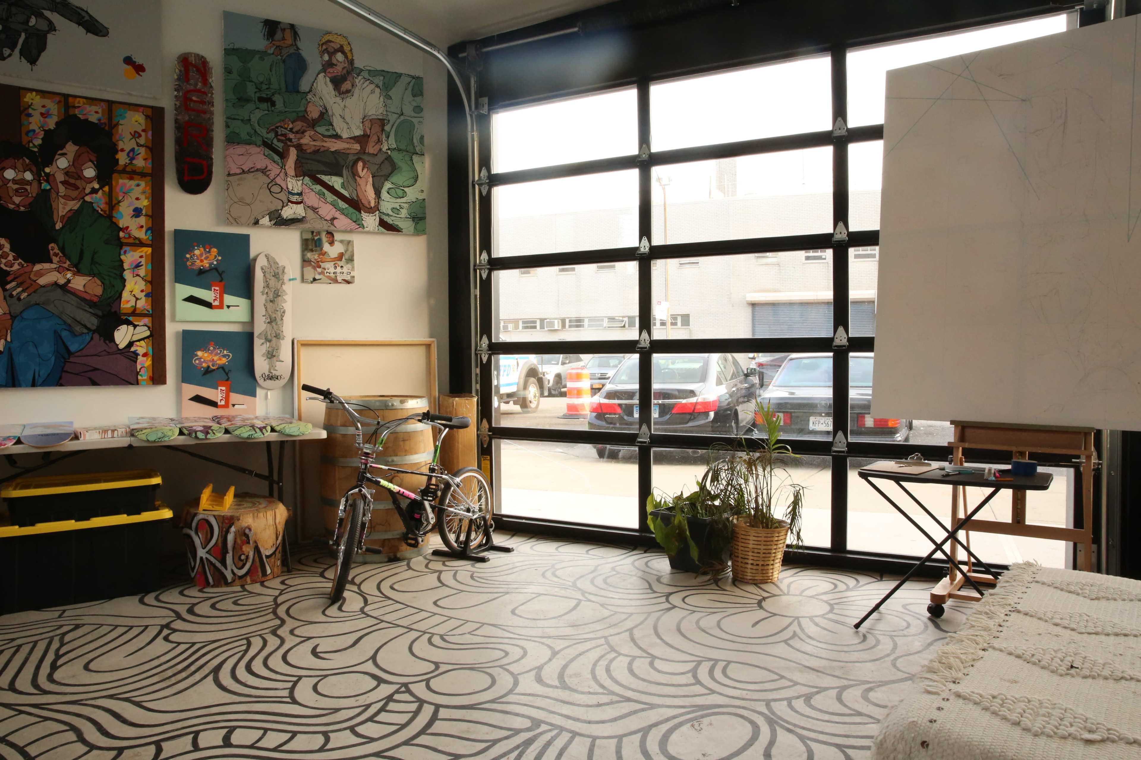 An artist's studio with a large garage door, artwork on the walls, a bicycle, a blank easel, and a patterned floor.