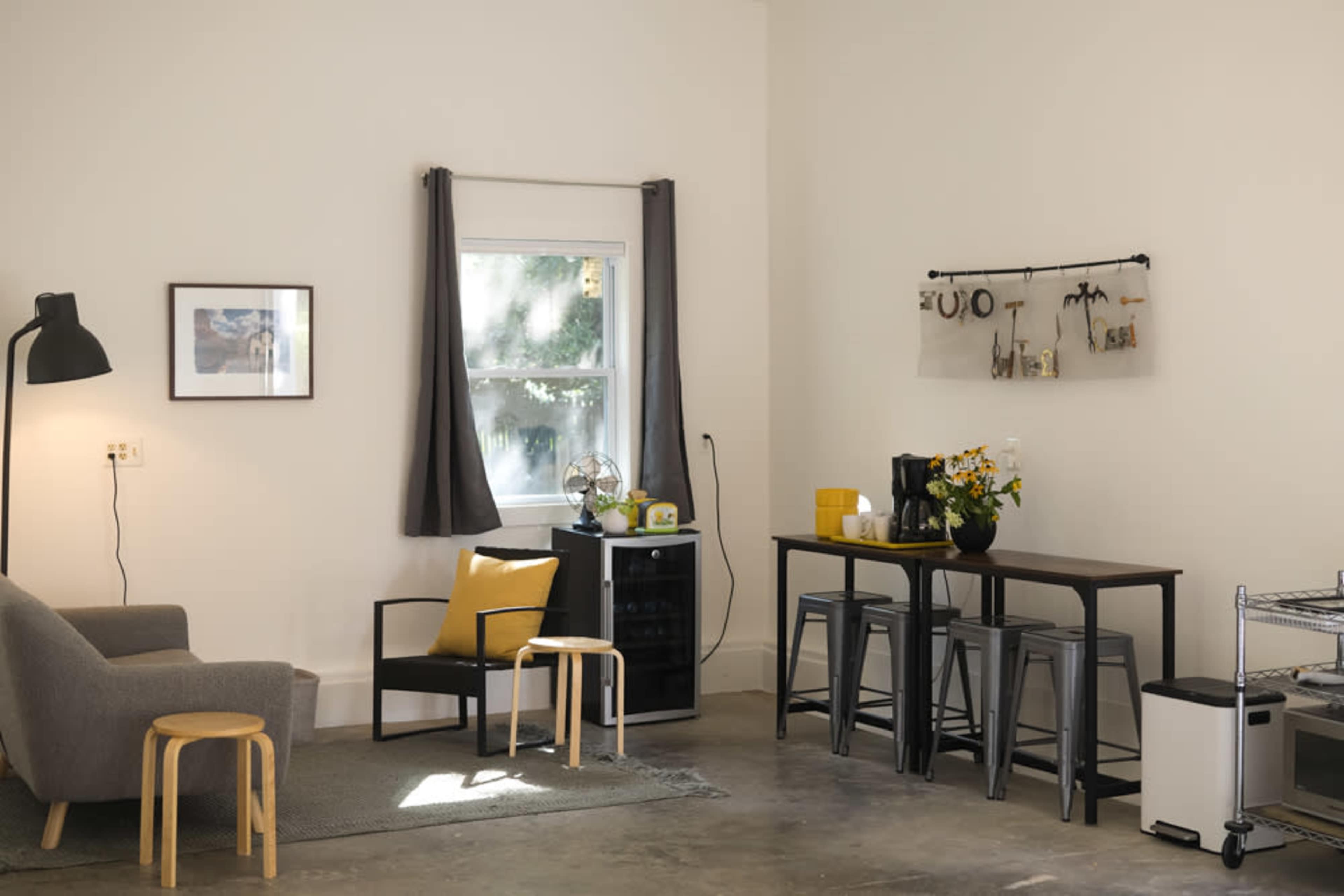 The image shows a modern living space with a gray sofa, a coffee table, and a dining area featuring tall stools around a table, complemented by a window and decor items on the wall.