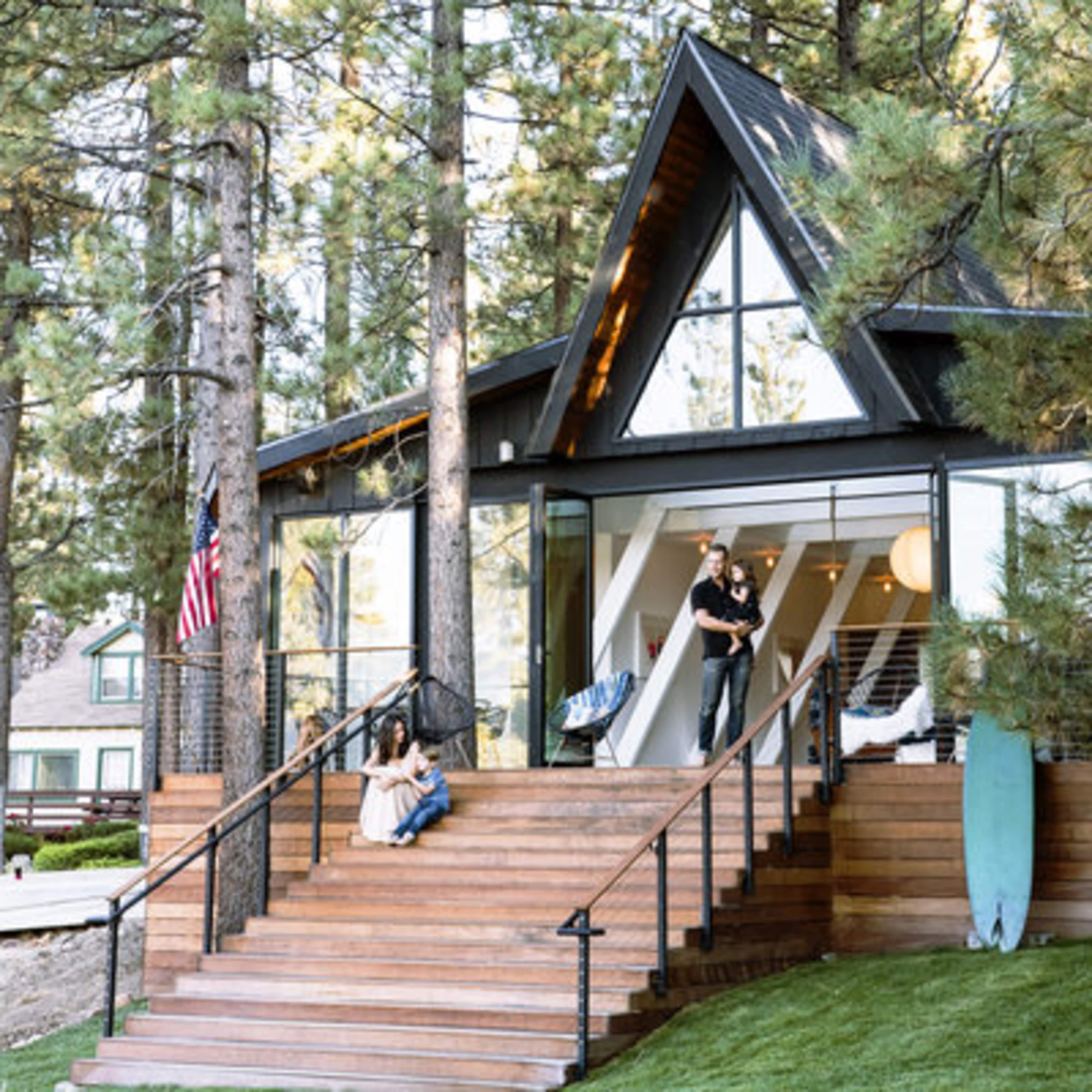 A modern A-frame house with large windows is surrounded by tall trees, featuring a wooden staircase leading to a porch where two people are engaged in conversation.