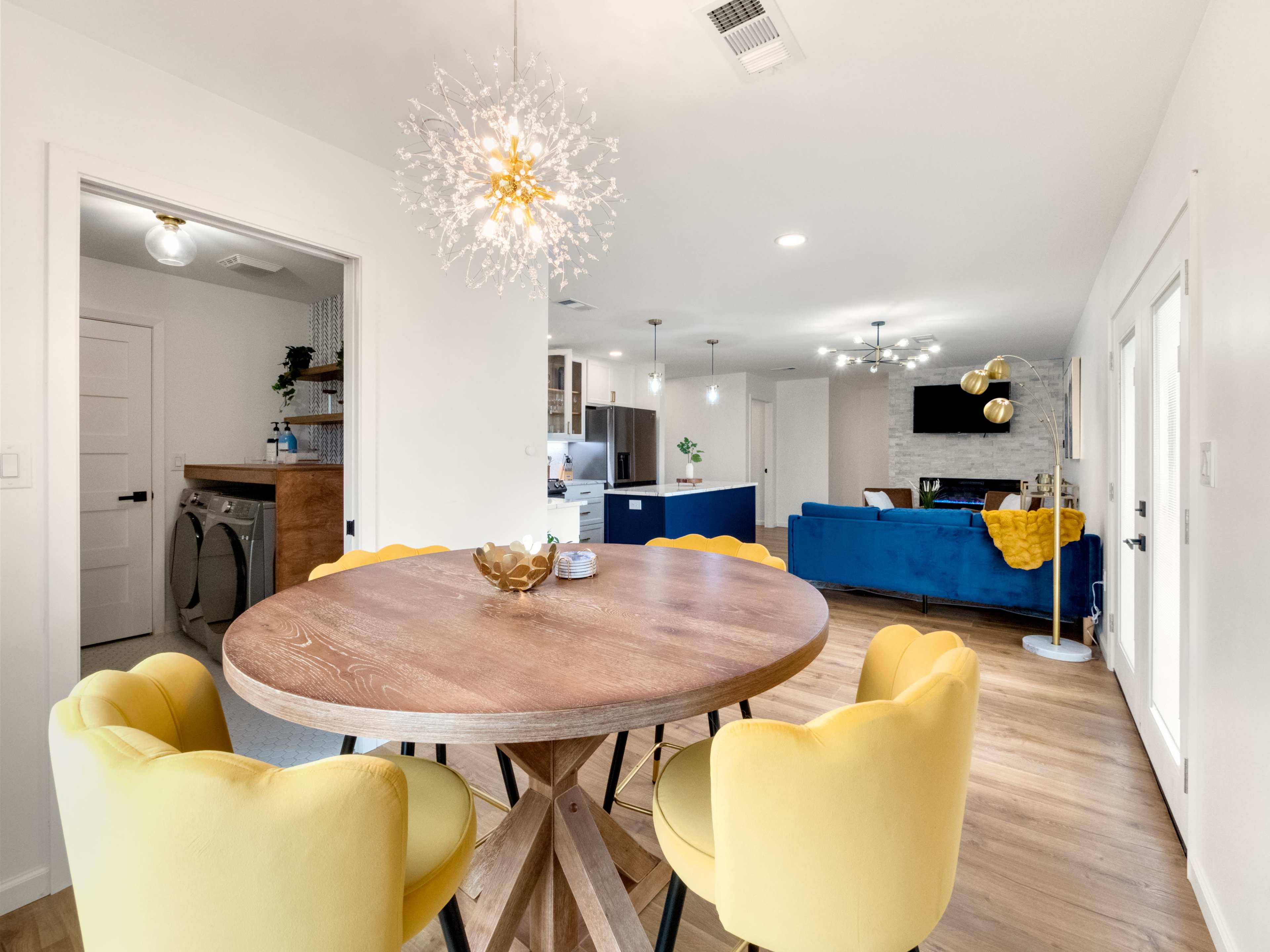 A round wooden dining table with yellow chairs is positioned in a brightly lit open-concept living space featuring a kitchen and a blue sofa.
