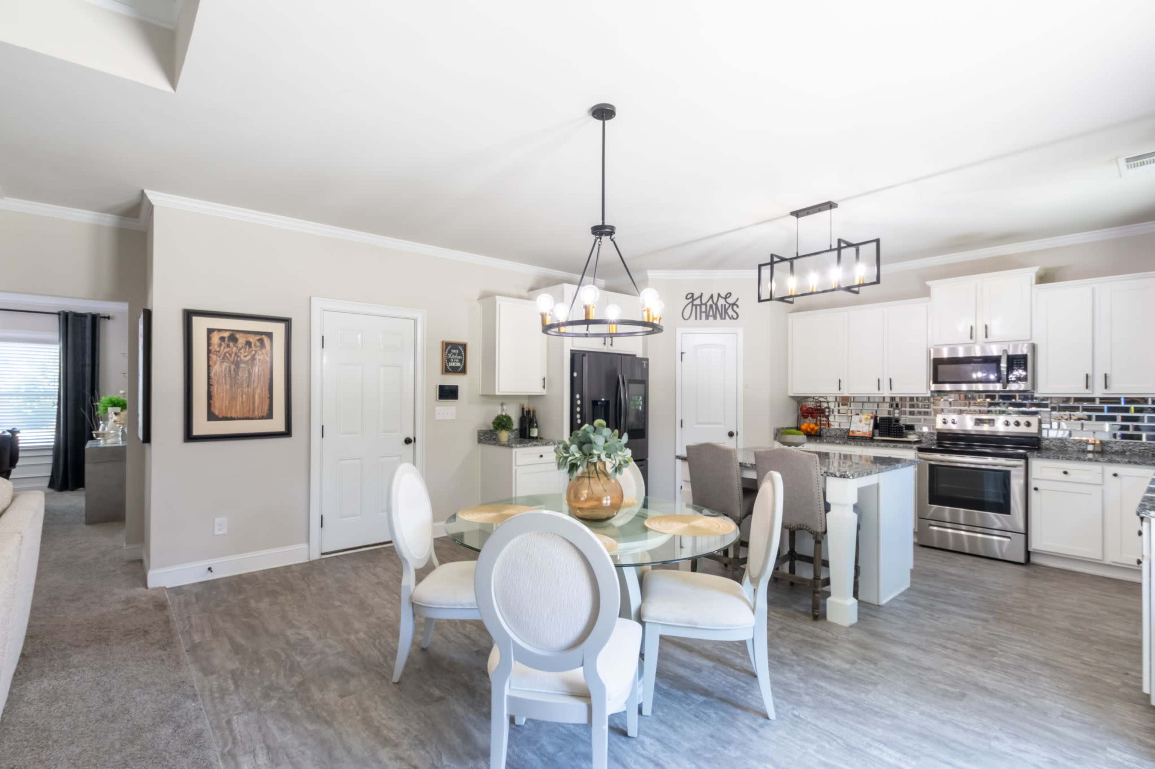 The image shows a modern kitchen and dining area with a circular glass table surrounded by upholstered chairs, a dark refrigerator, and white cabinetry complemented by gray countertops and tiled backsplash.