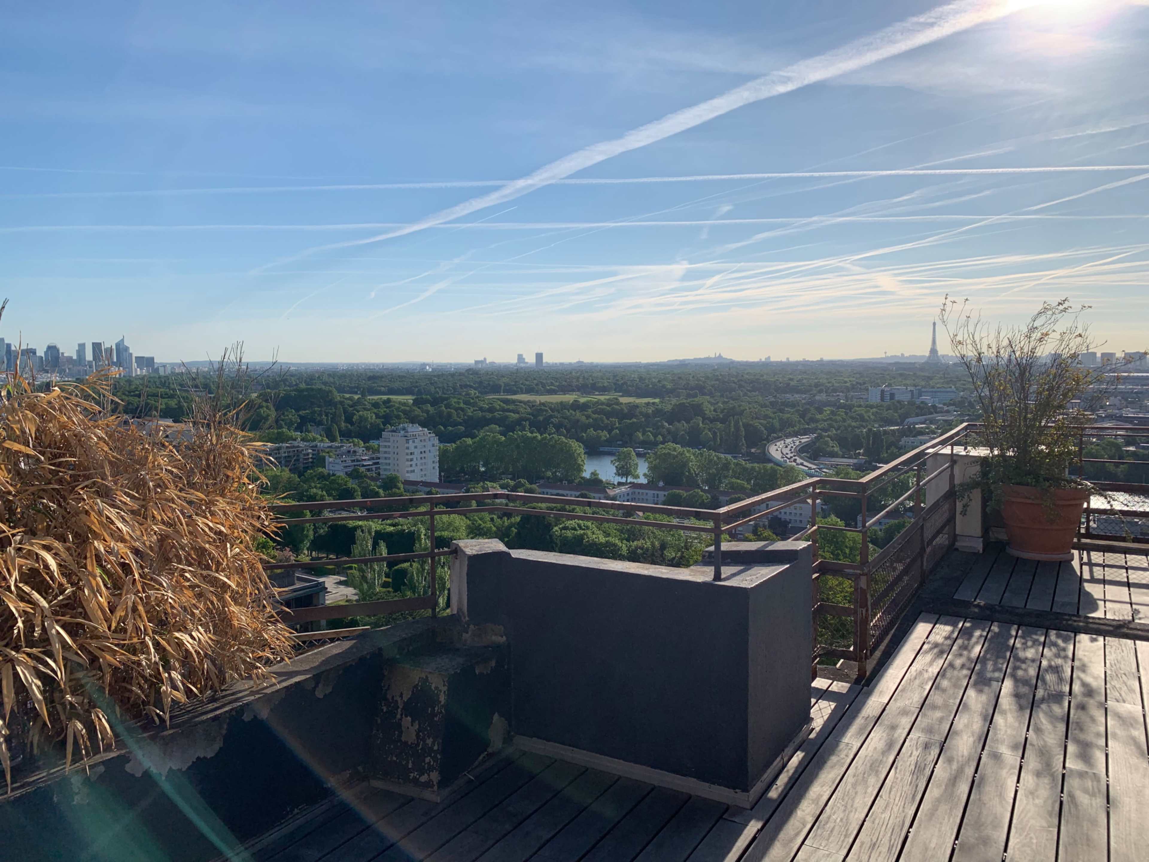 A rooftop terrace offers a panoramic view of a green landscape, city skyline, and a clear blue sky with wispy clouds.