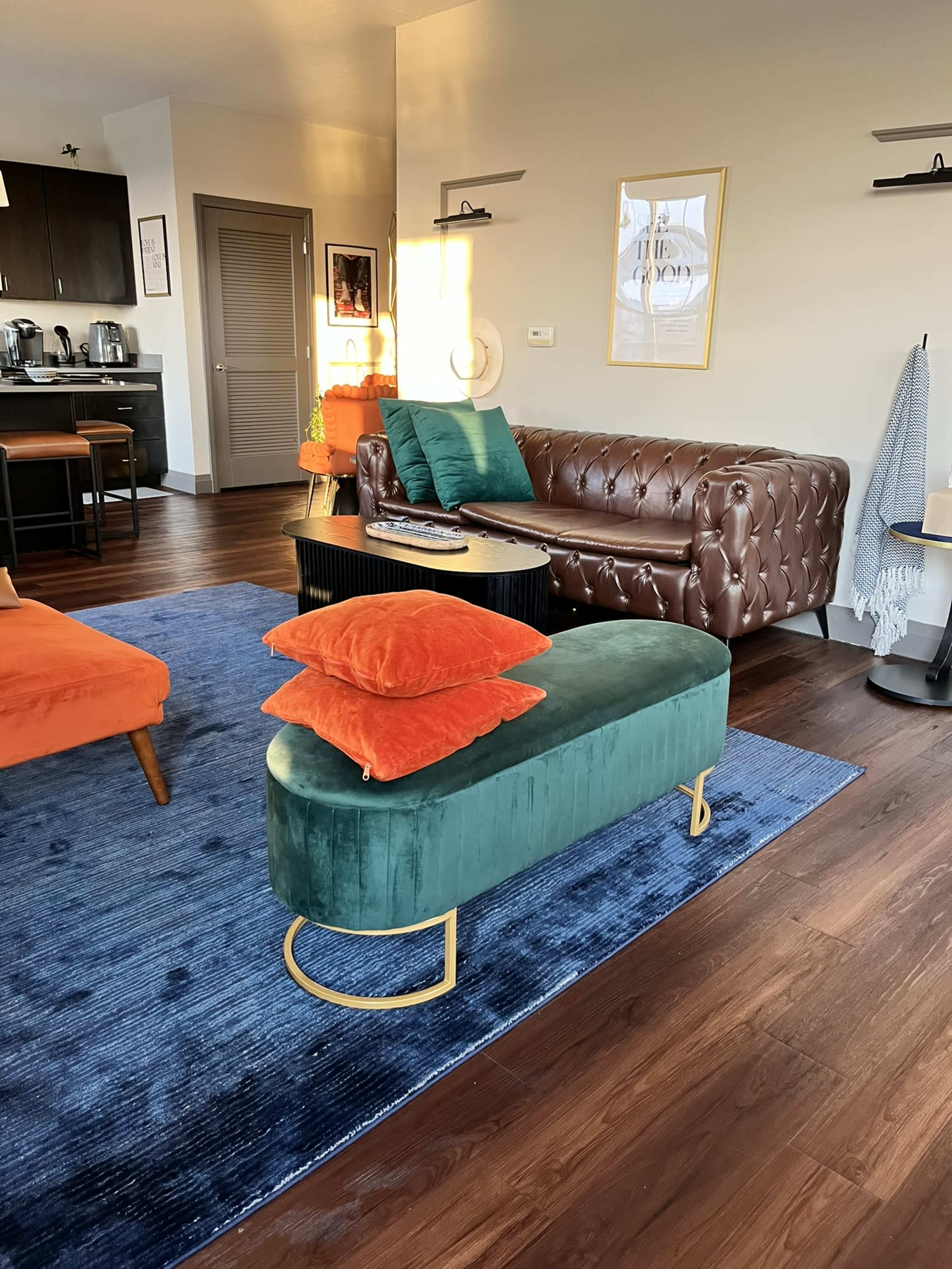 The image shows a modern living room with a brown leather sofa, a green velvet bench, orange pillows, and kitchen appliances visible in the background.