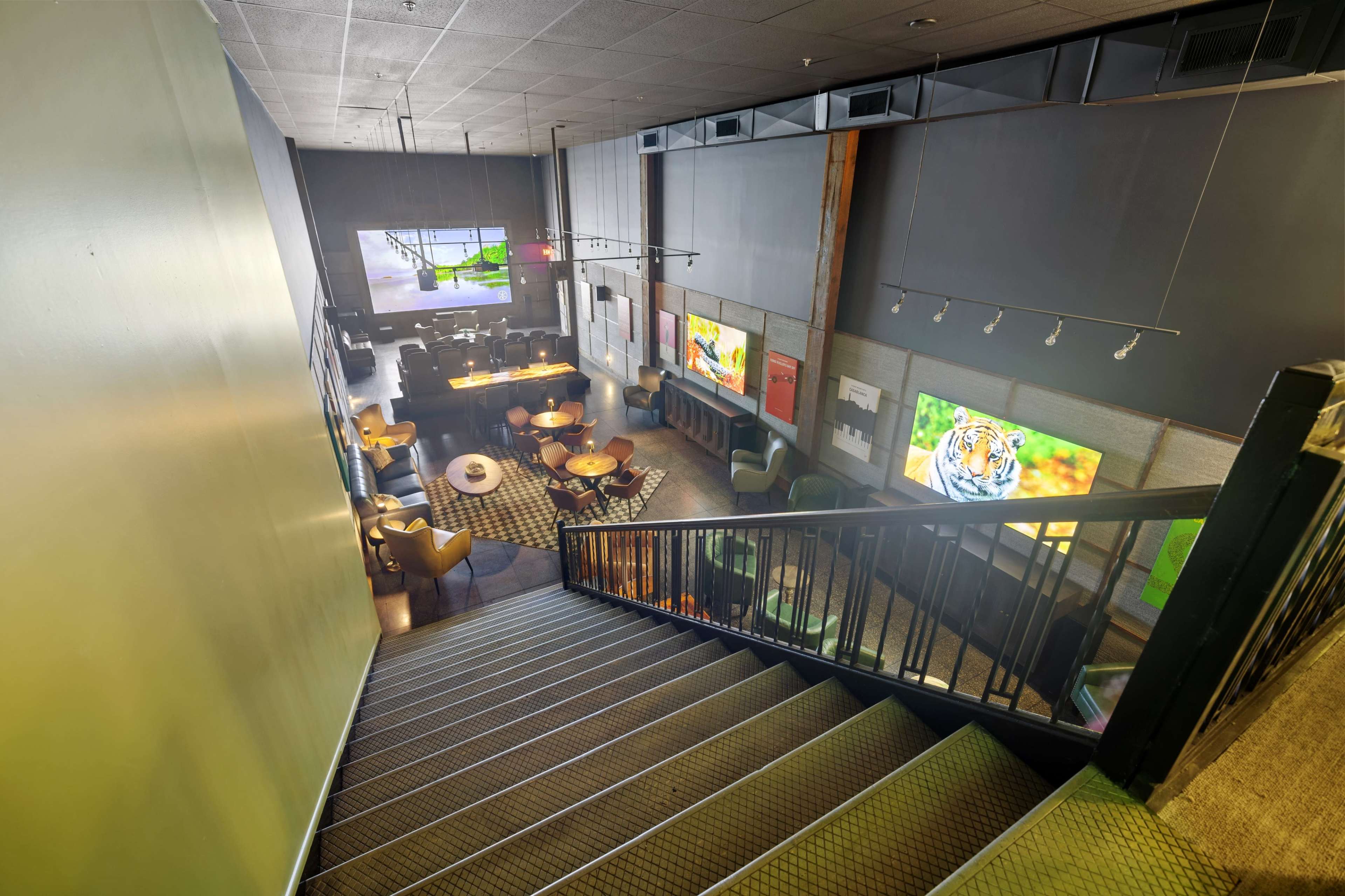 The image shows a staircase leading down to a modern, partially lit lounge area with tables, chairs, and large wall-mounted screens displaying images.