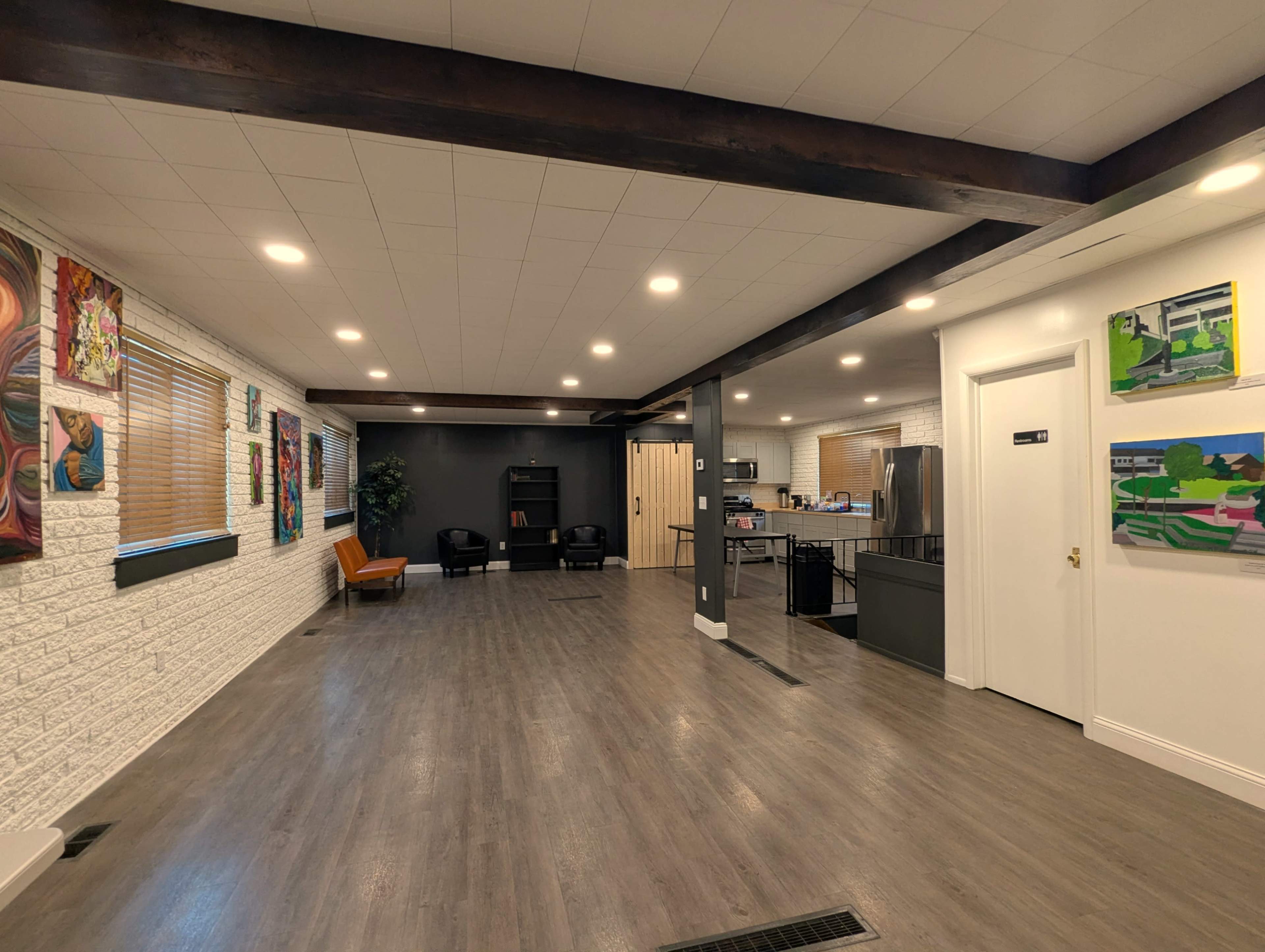 The image shows a spacious, modern interior with a wooden beam ceiling, artwork on the walls, and distinct areas for seating and kitchen facilities.