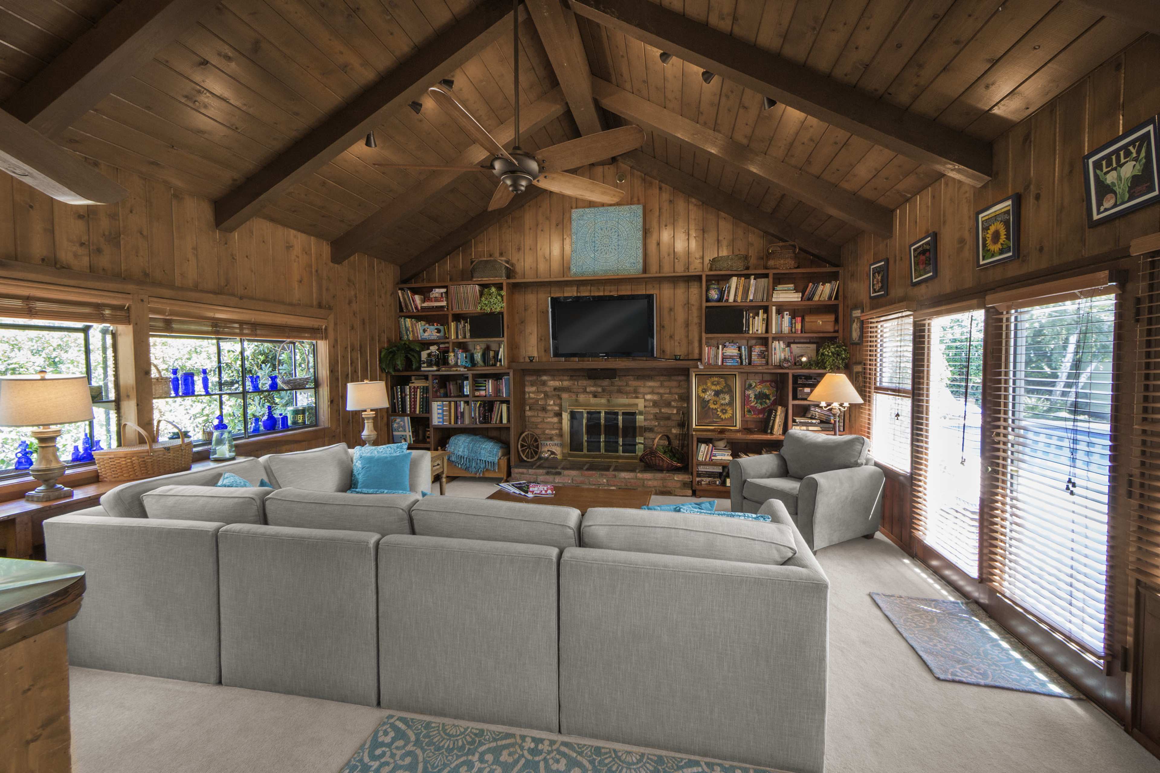 The image shows a rustic living room with wooden walls and a large couch facing a brick fireplace and a television, flanked by bookshelves and large windows.