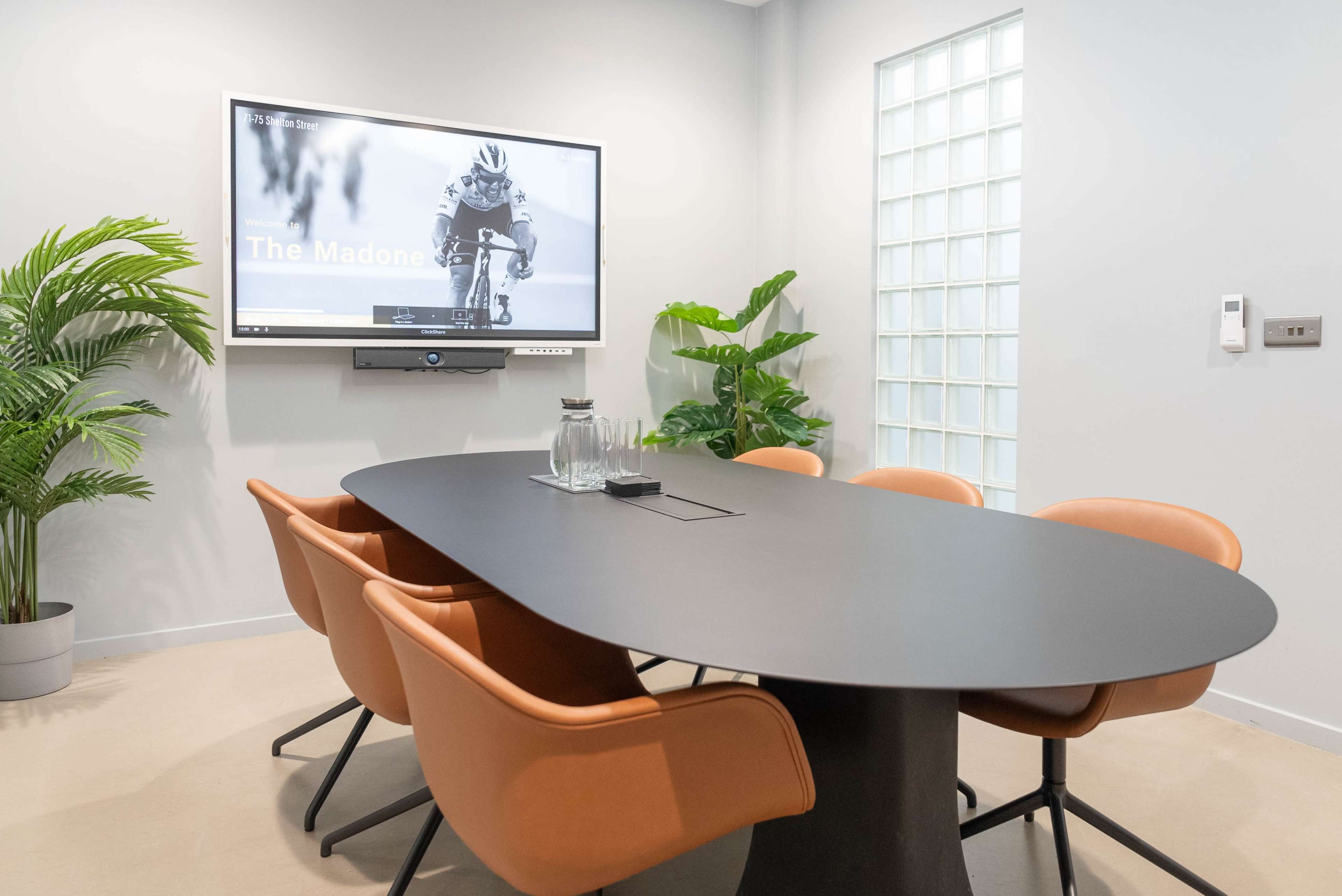 A modern conference room features a black oval table surrounded by orange chairs, a large screen on the wall displaying an image of a cyclist, and potted plants in the corners.