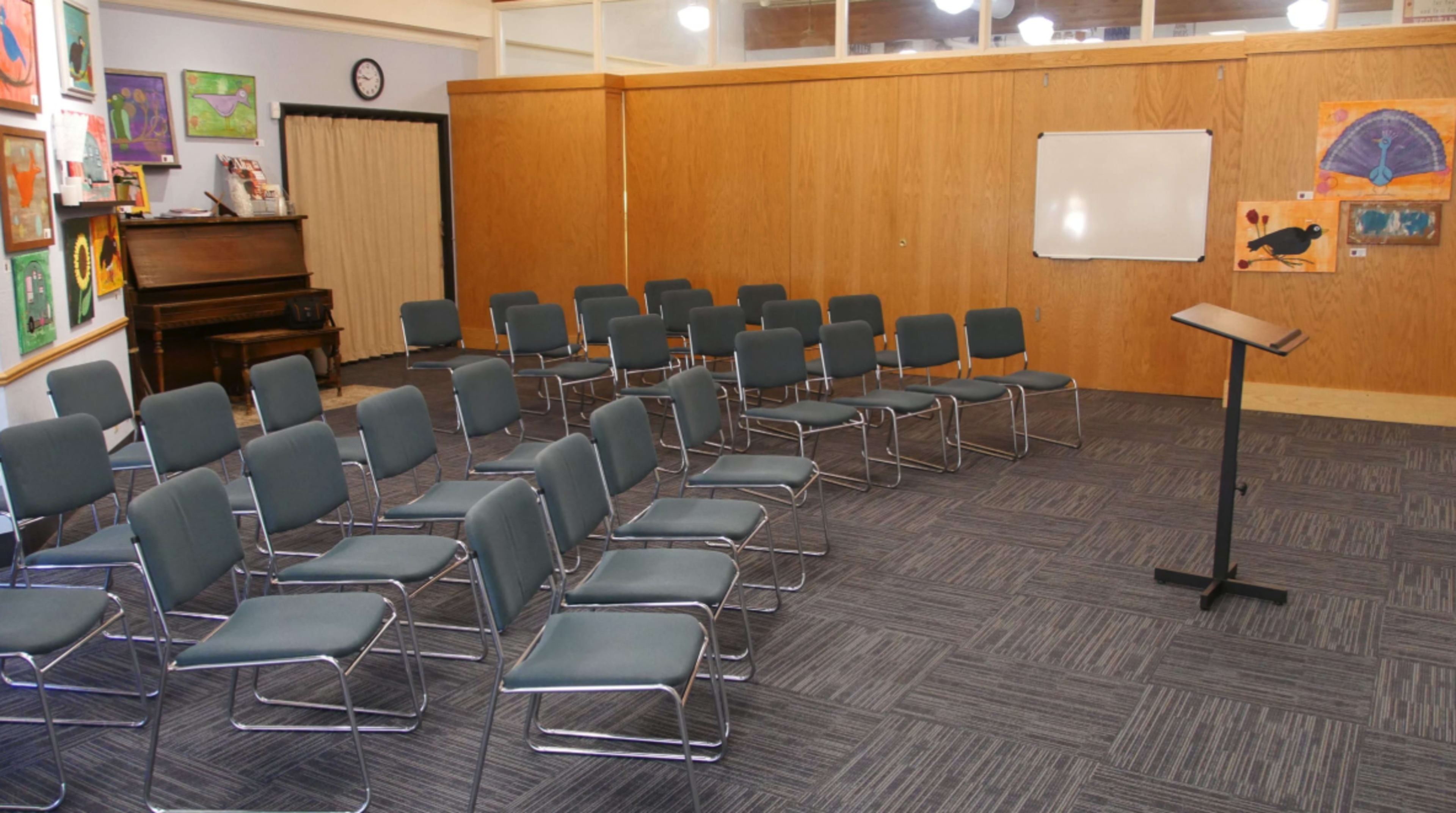 A room with several rows of metal-framed chairs arranged facing a podium, with a piano and colorful artwork displayed on the walls.