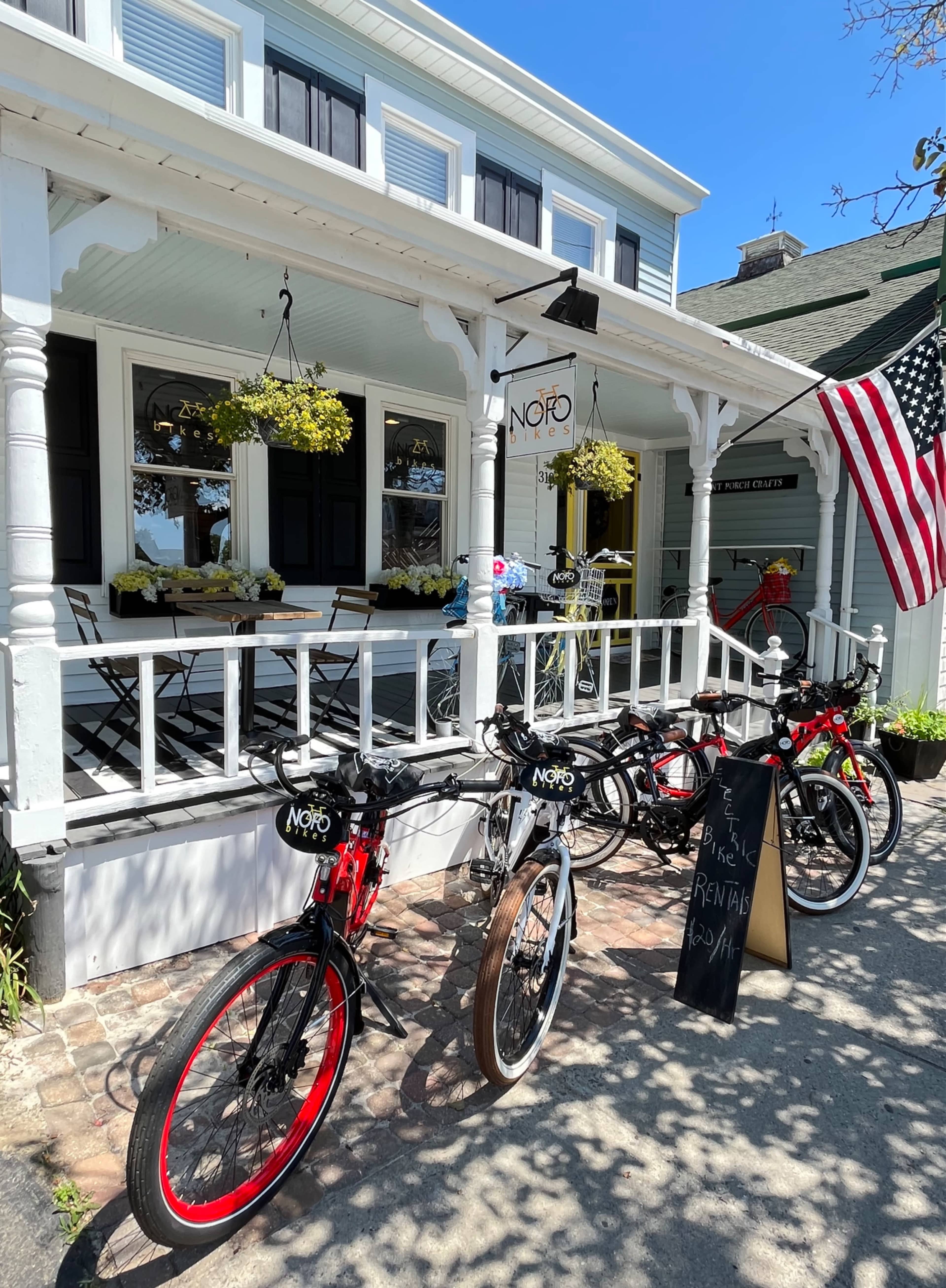 A row of bicycles is parked in front of a building with a sign that reads "NOFO" and hanging flower planters, alongside an American flag.
