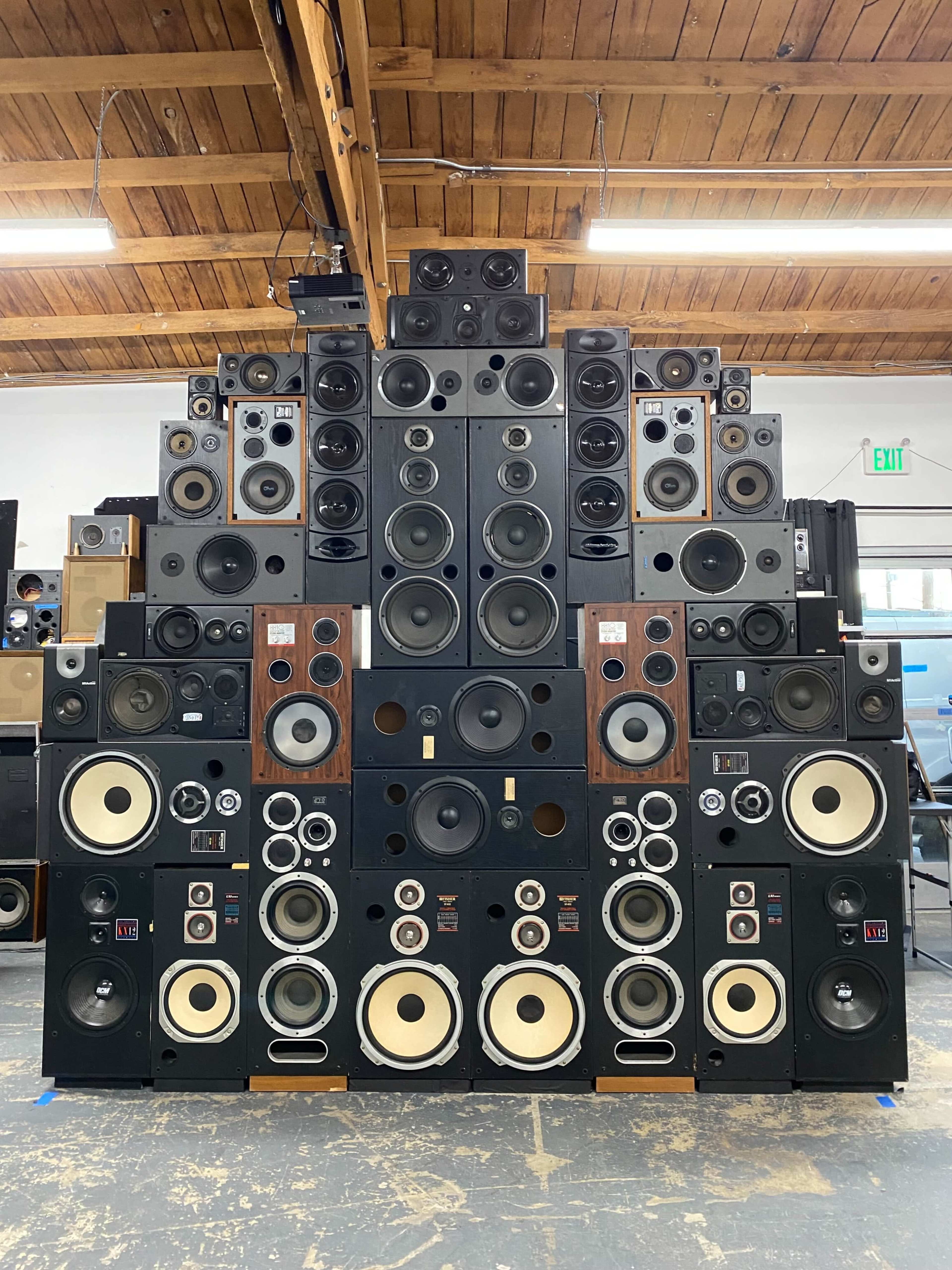 A large arrangement of various speaker cabinets is displayed against a wooden ceiling in a spacious room.