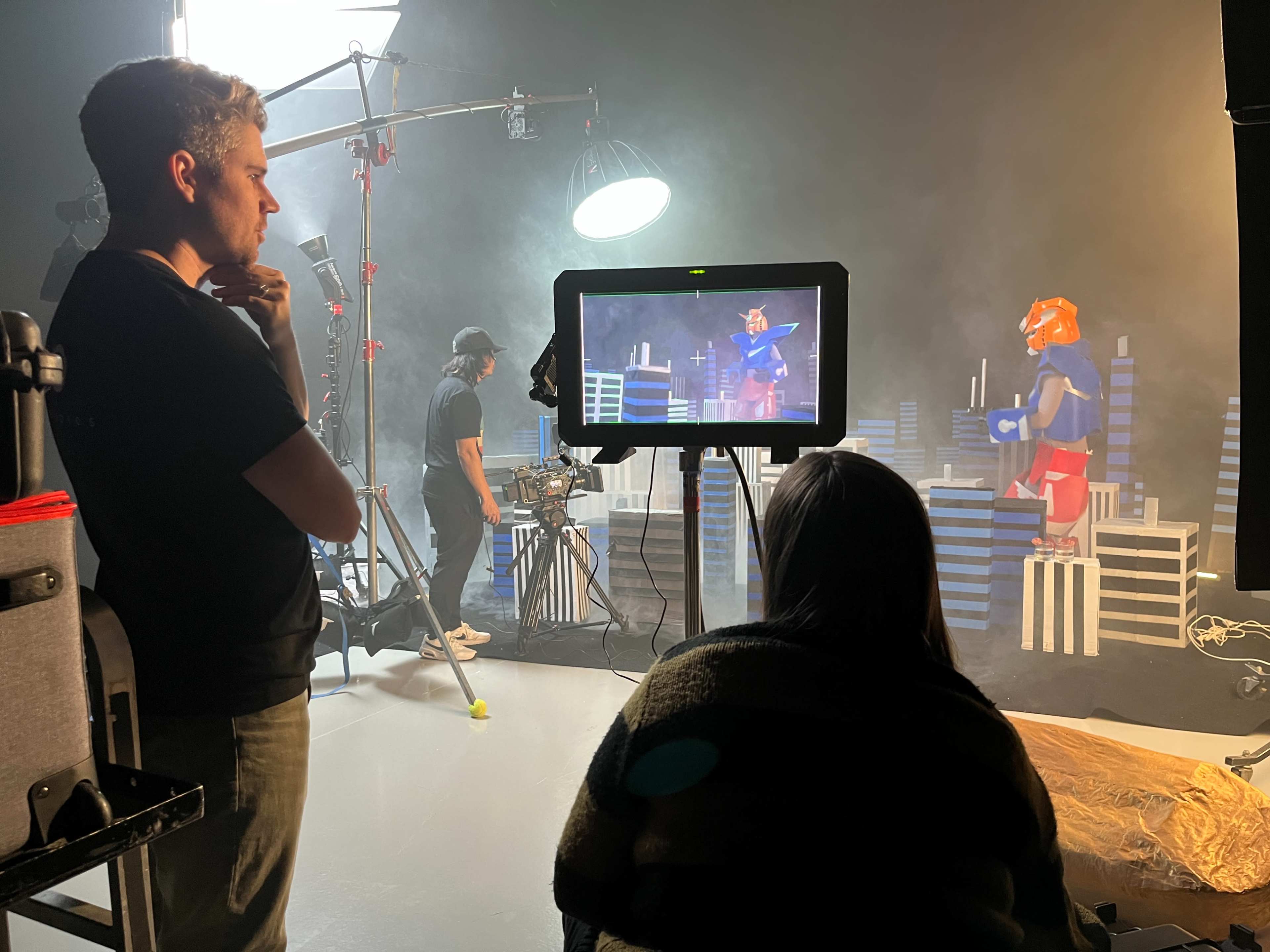 A crew works on a film set, with one person monitoring a screen while another adjusts equipment, and a character in costume poses against a backdrop of props.