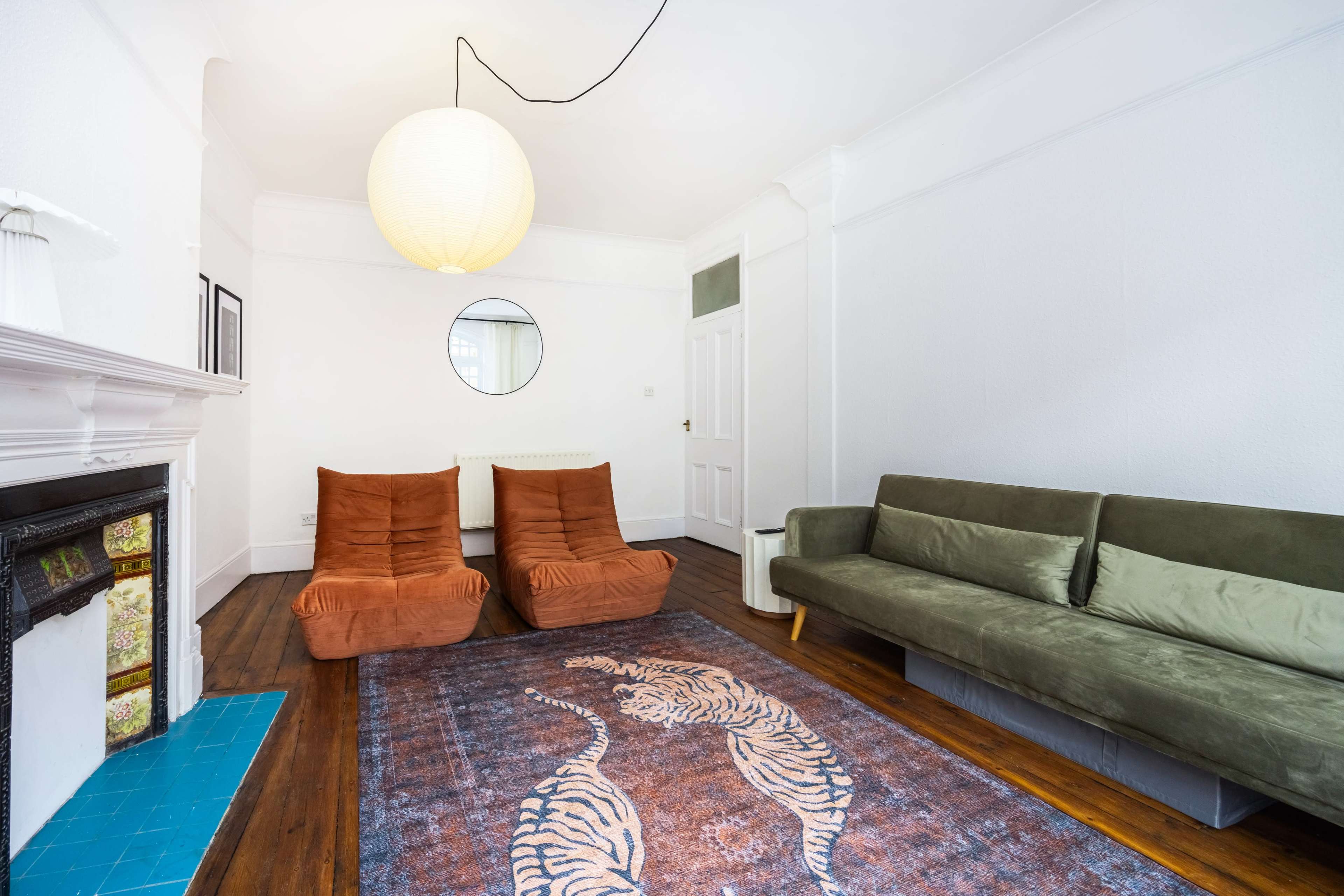 The image shows a minimalist living room with two orange armchairs, a green sofa, and a round mirror on the white wall, featuring wooden flooring and a patterned rug.