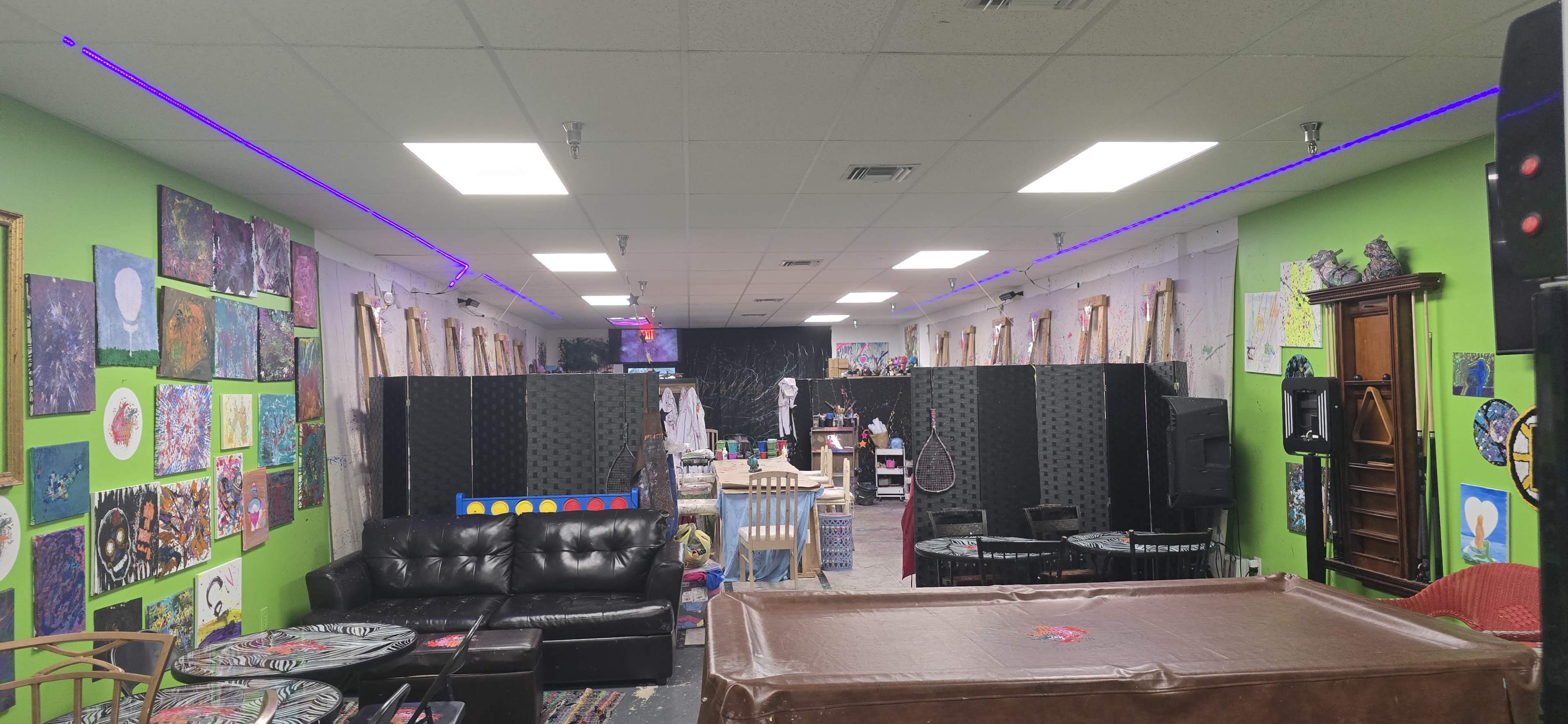 The image shows a colorful art studio with green walls, several artworks displayed, a black couch, and decorative lighting.