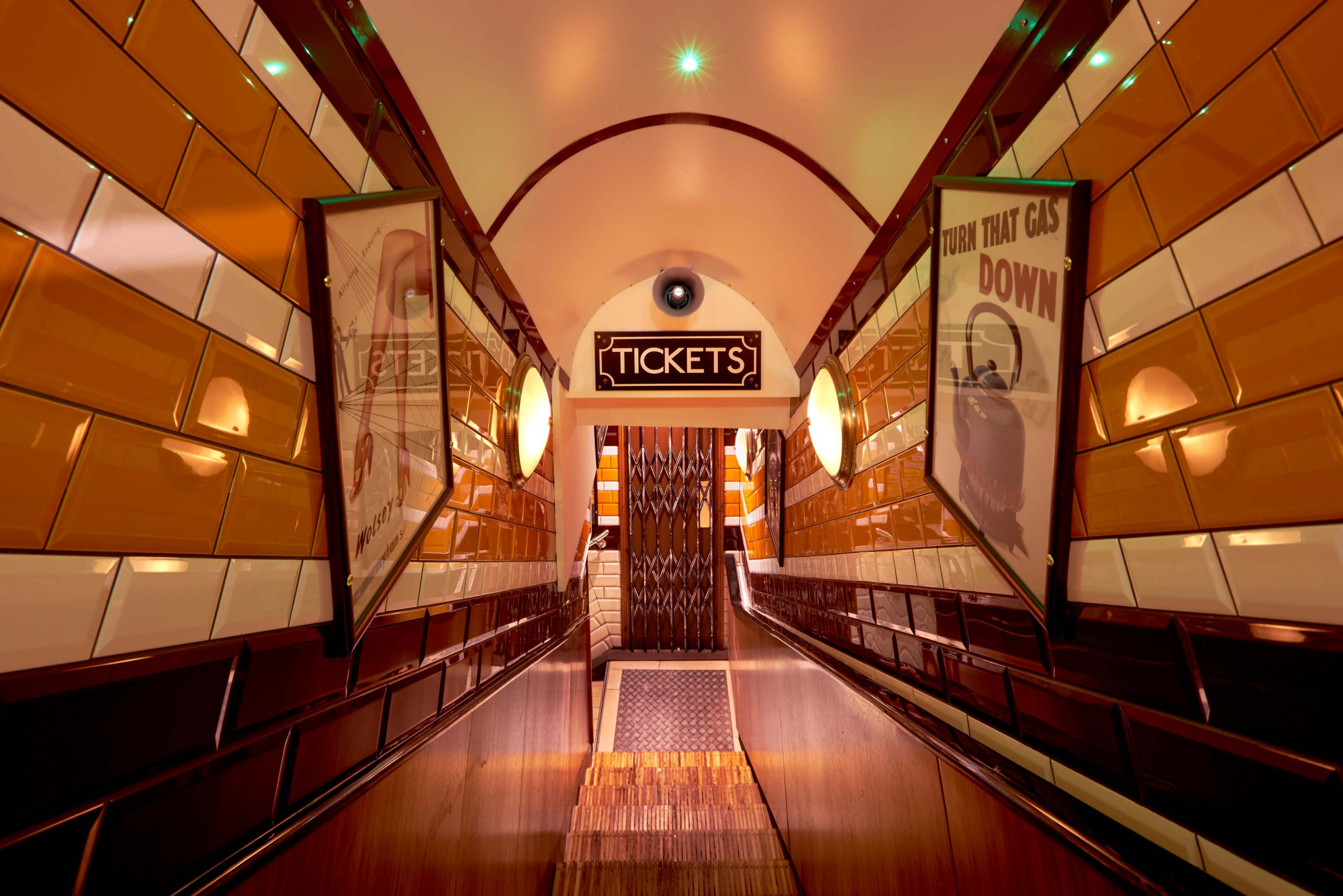 A downward sloping corridor lined with glossy orange and white tiles, leading to a "TICKETS" sign at the entrance of an event space or venue.