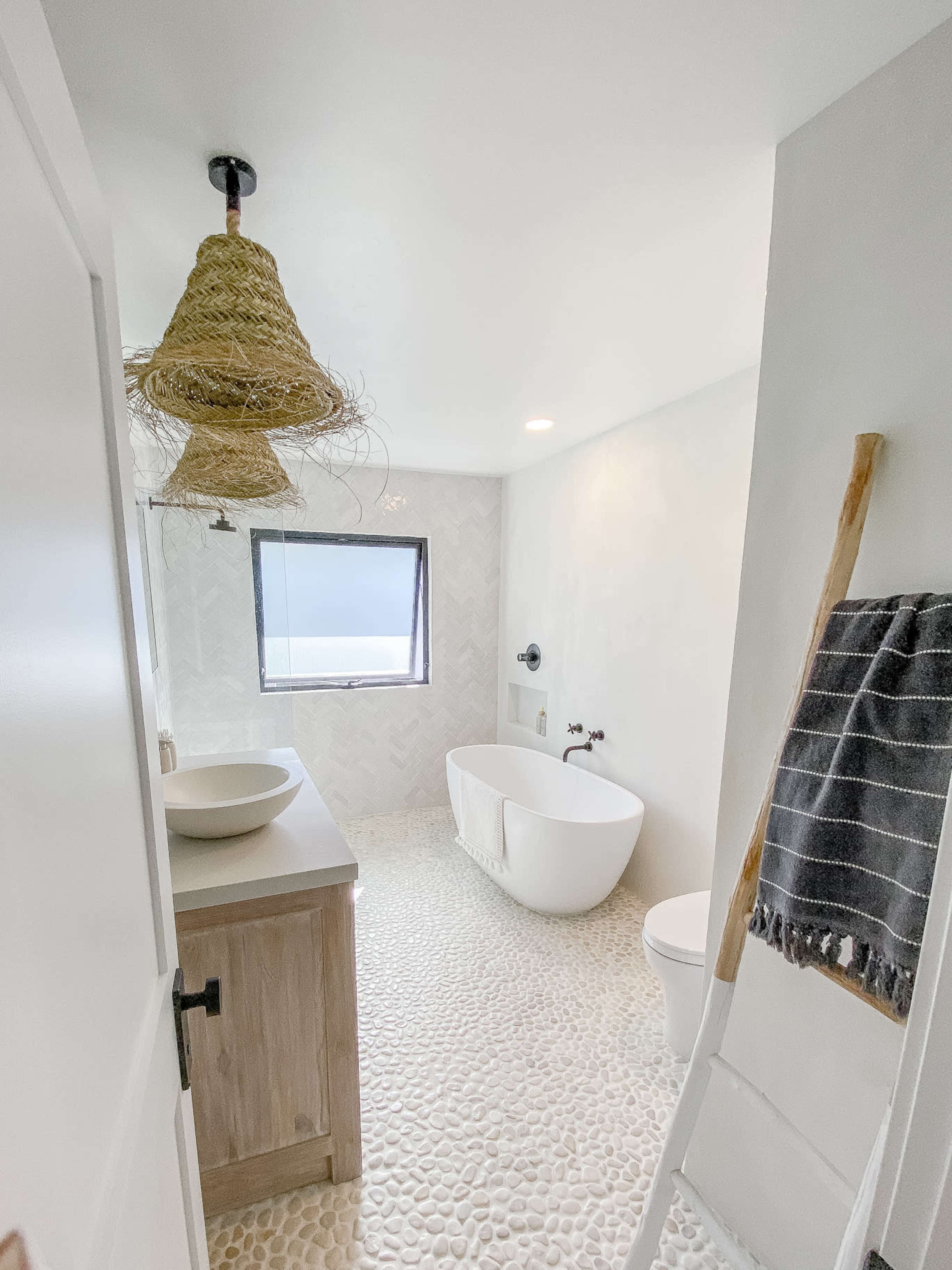 A modern bathroom with a freestanding bathtub, a wooden vanity, a pebble stone floor, and a large window.