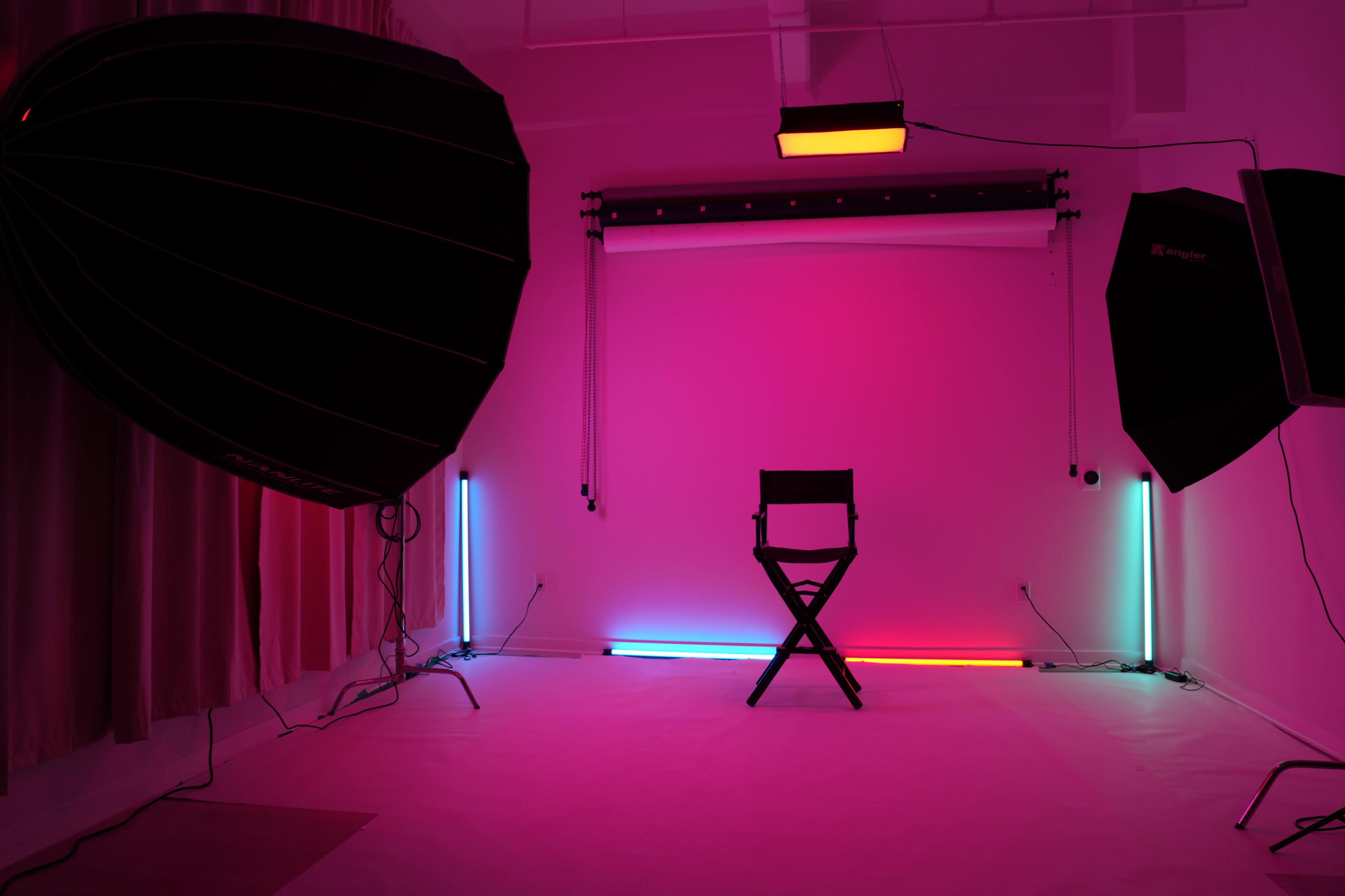 The image shows a photography studio with pink lighting, featuring a director's chair in the center and various softbox lights positioned around it.