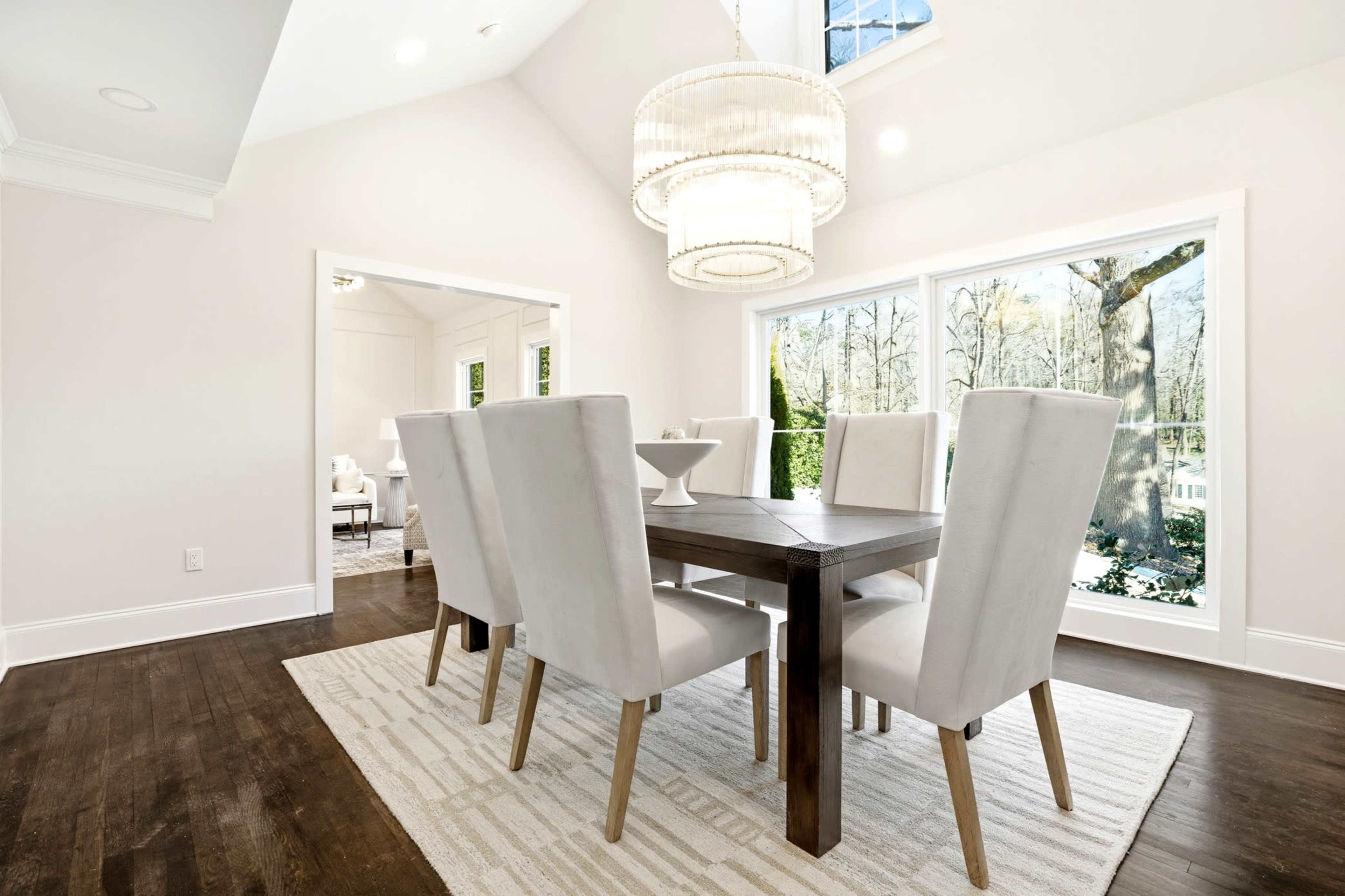 A modern dining room features a rectangular wooden table with six upholstered chairs, illuminated by a large chandelier and flanked by two large windows.