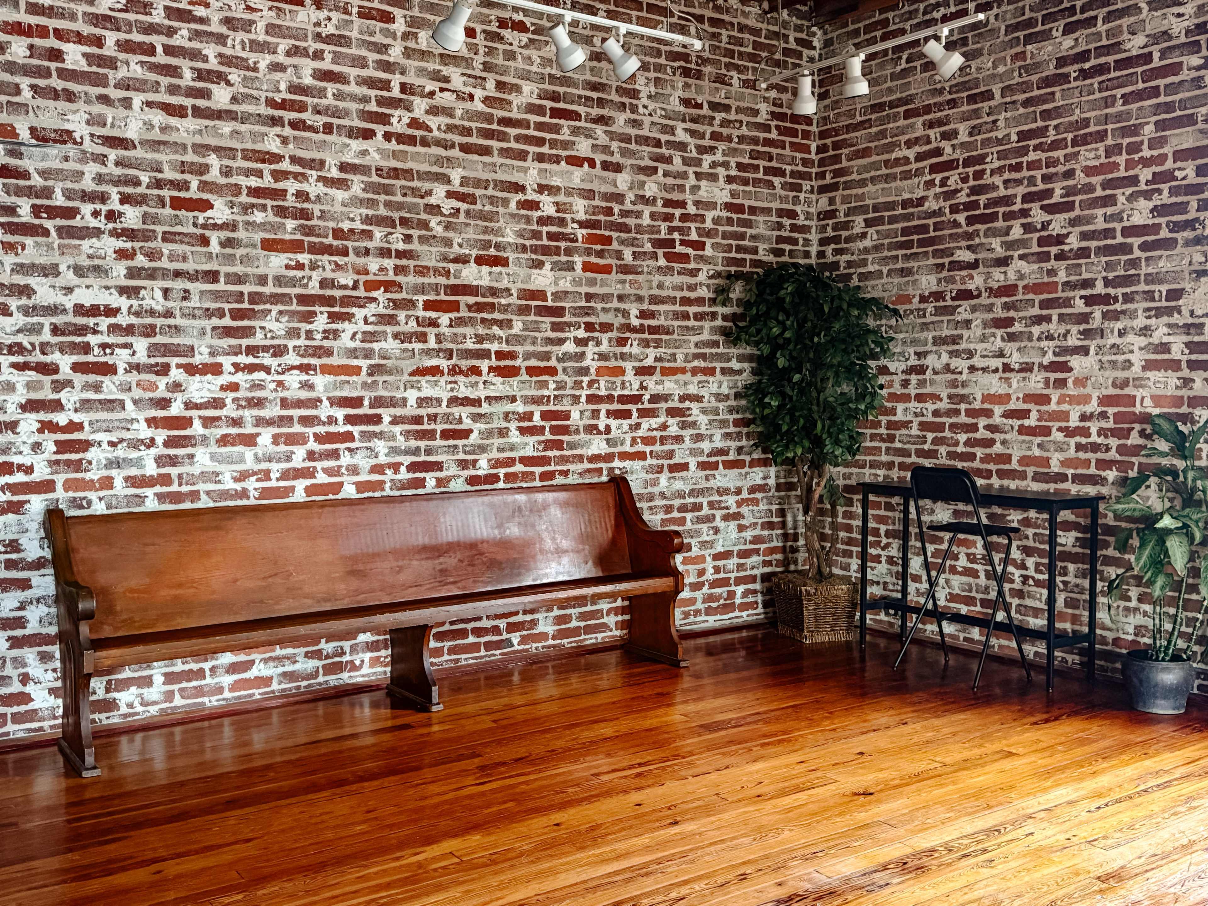 The image shows a room with a wooden bench against a brick wall, a small table with a chair beside it, and a potted plant in the corner.