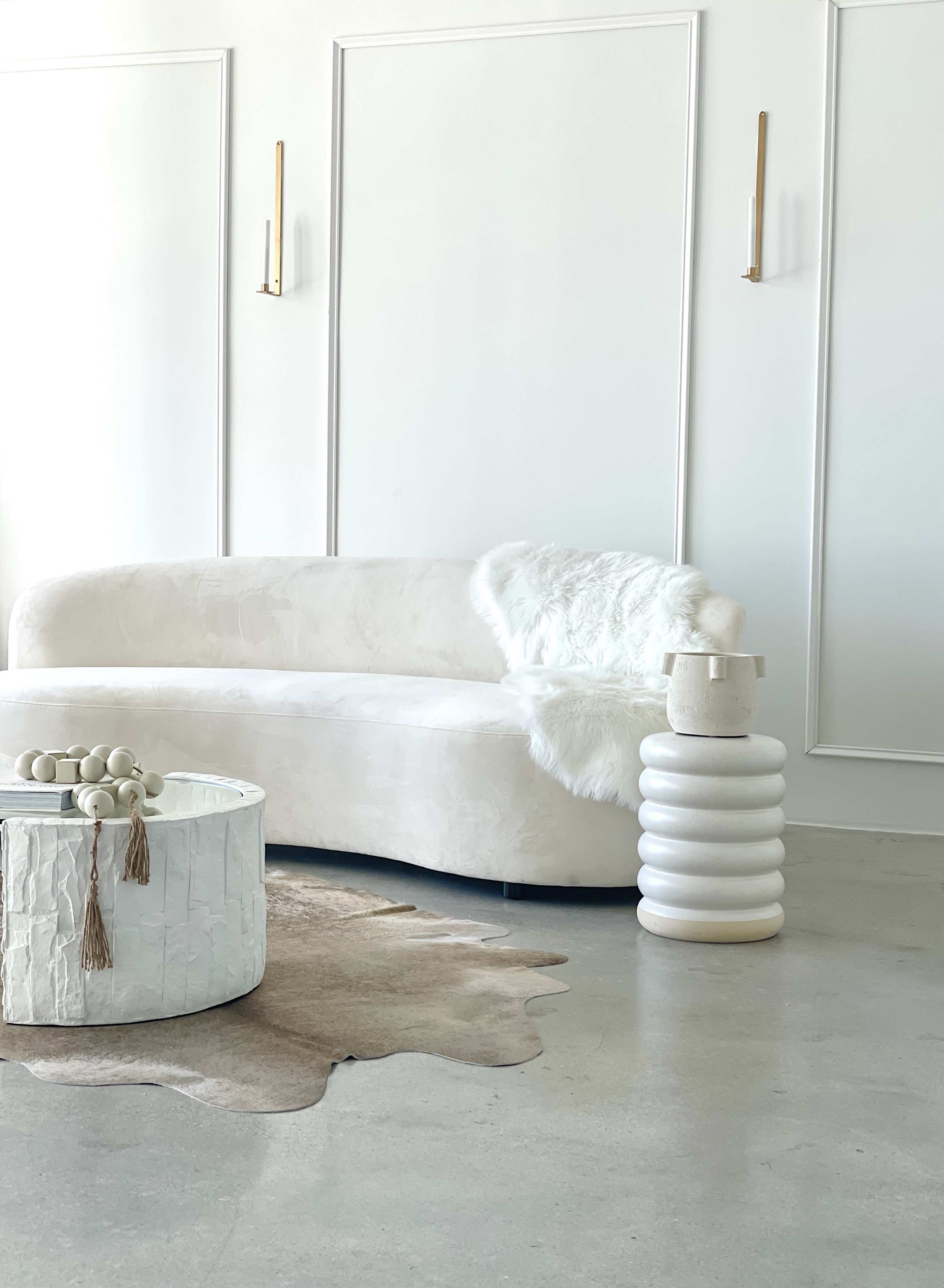 A modern living room features a white curved sofa, a round coffee table with a textured surface, and a decorative side table in a minimalist design, all set against a light-colored wall.