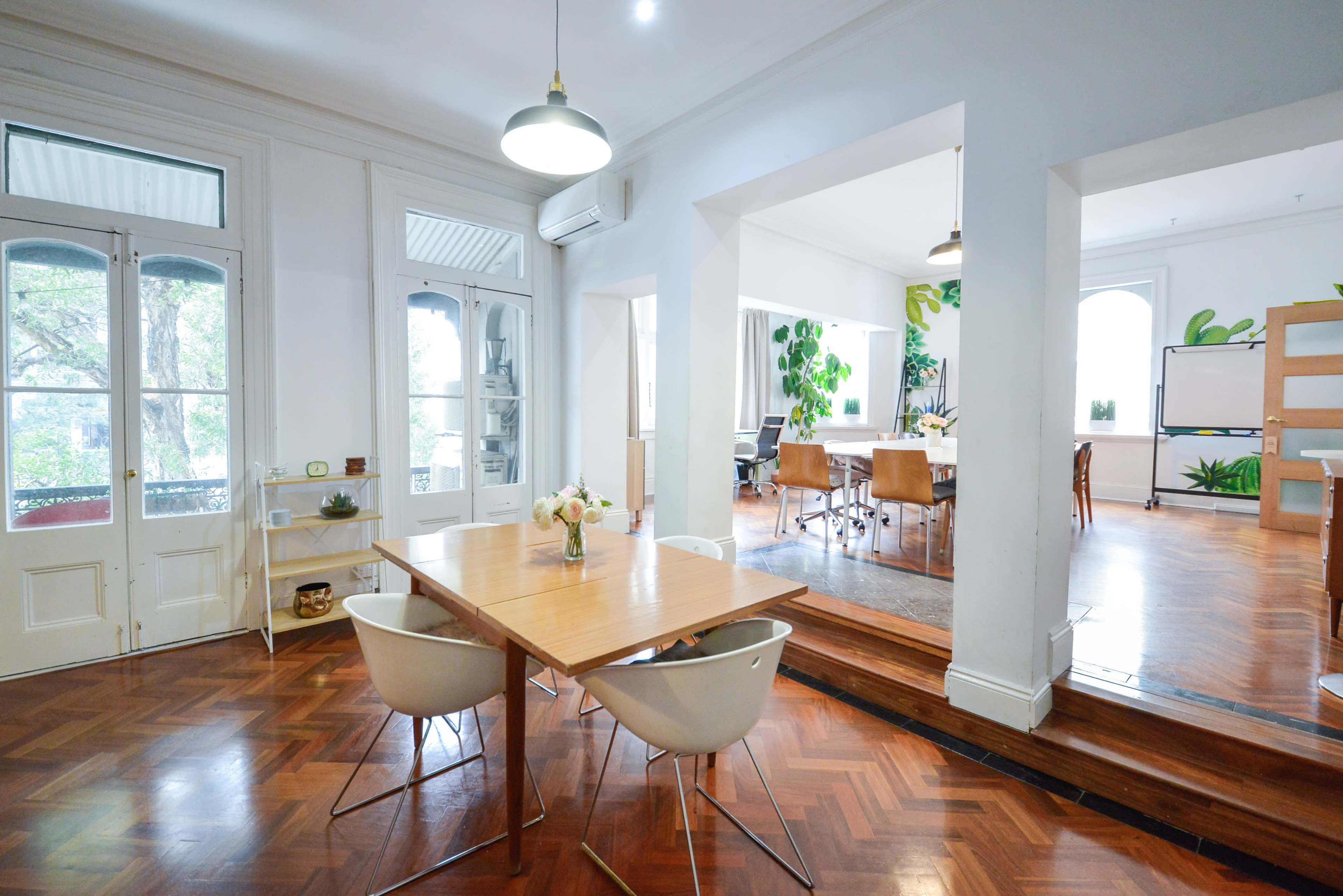 A bright, open dining area featuring a wooden table with white chairs, leading into a well-lit office space with plants and modern furniture.