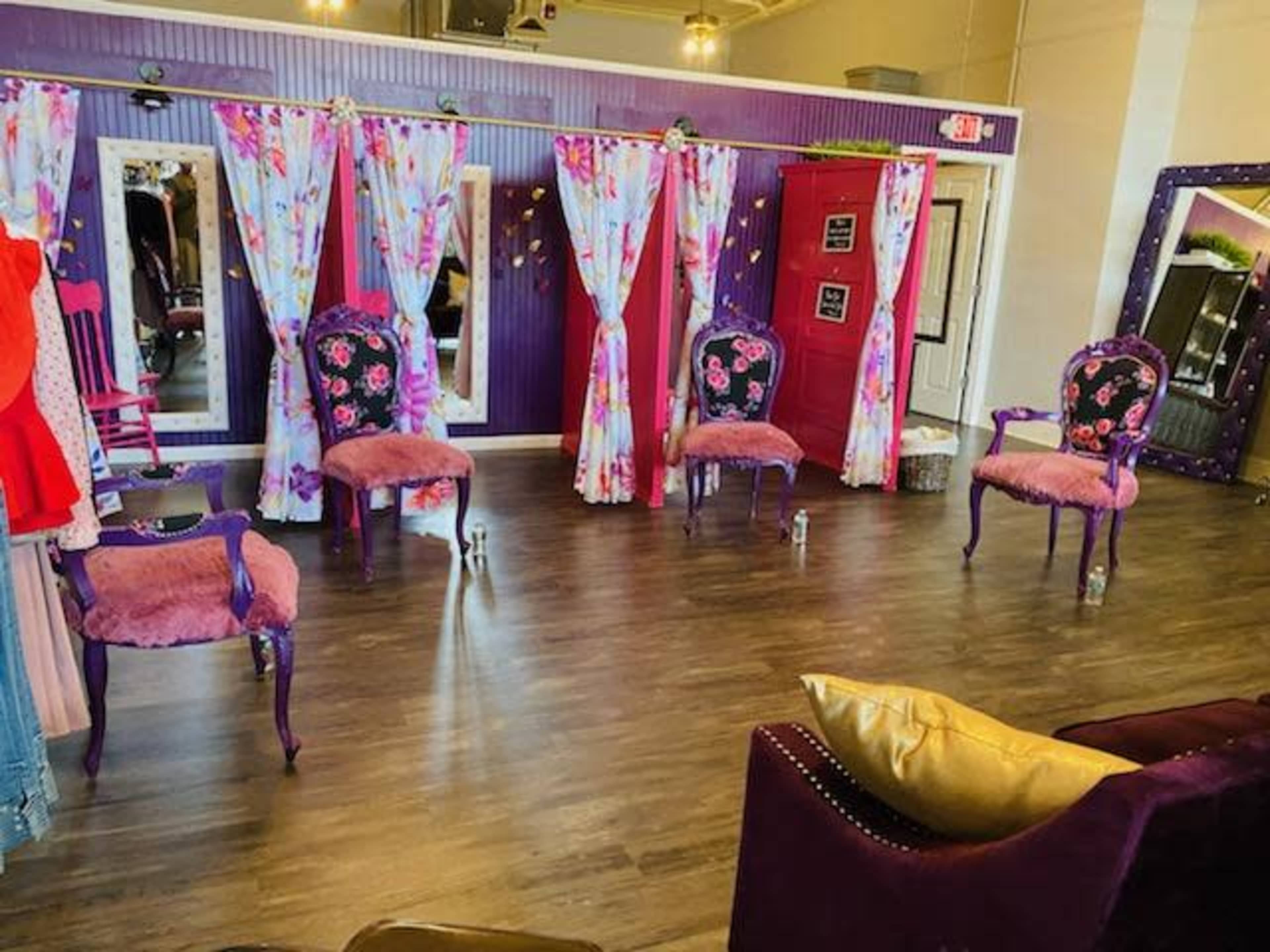 A bright boutique interior with floral curtains, three purple chairs with floral upholstery, and a mirrored wall.