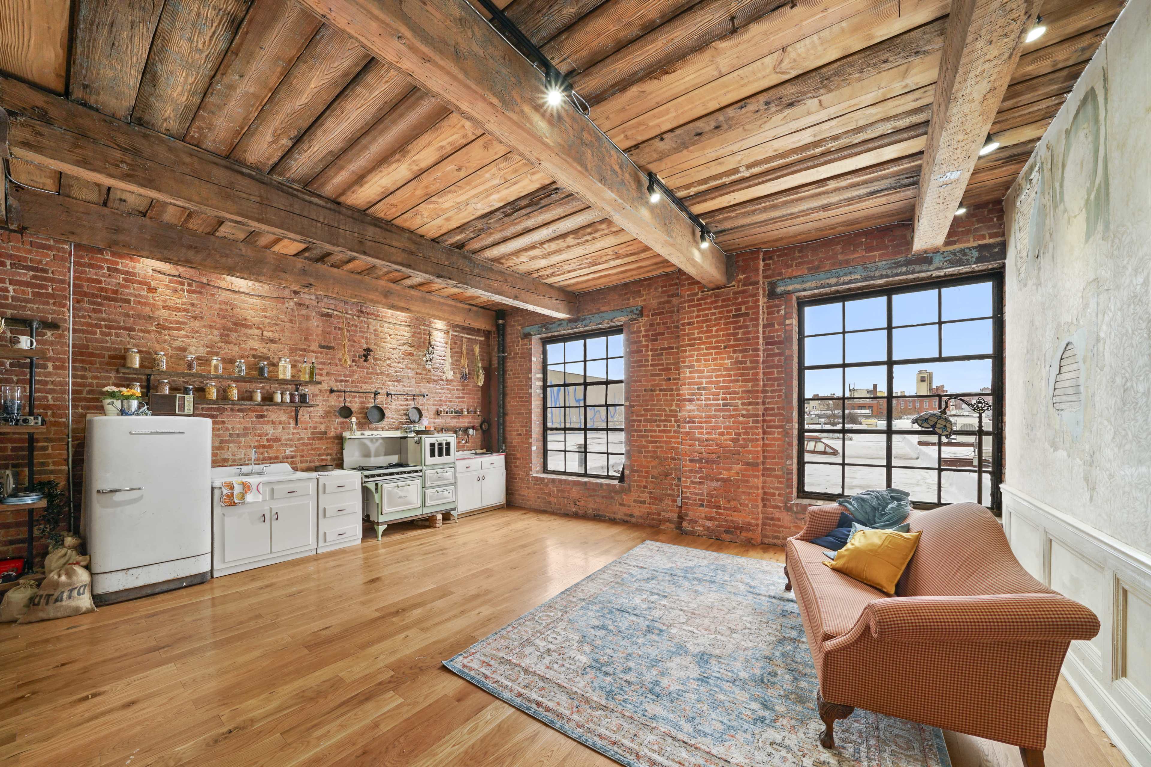 A spacious interior with exposed brick walls, wooden beams, and large windows, furnished with a vintage couch and a retro kitchen setup.