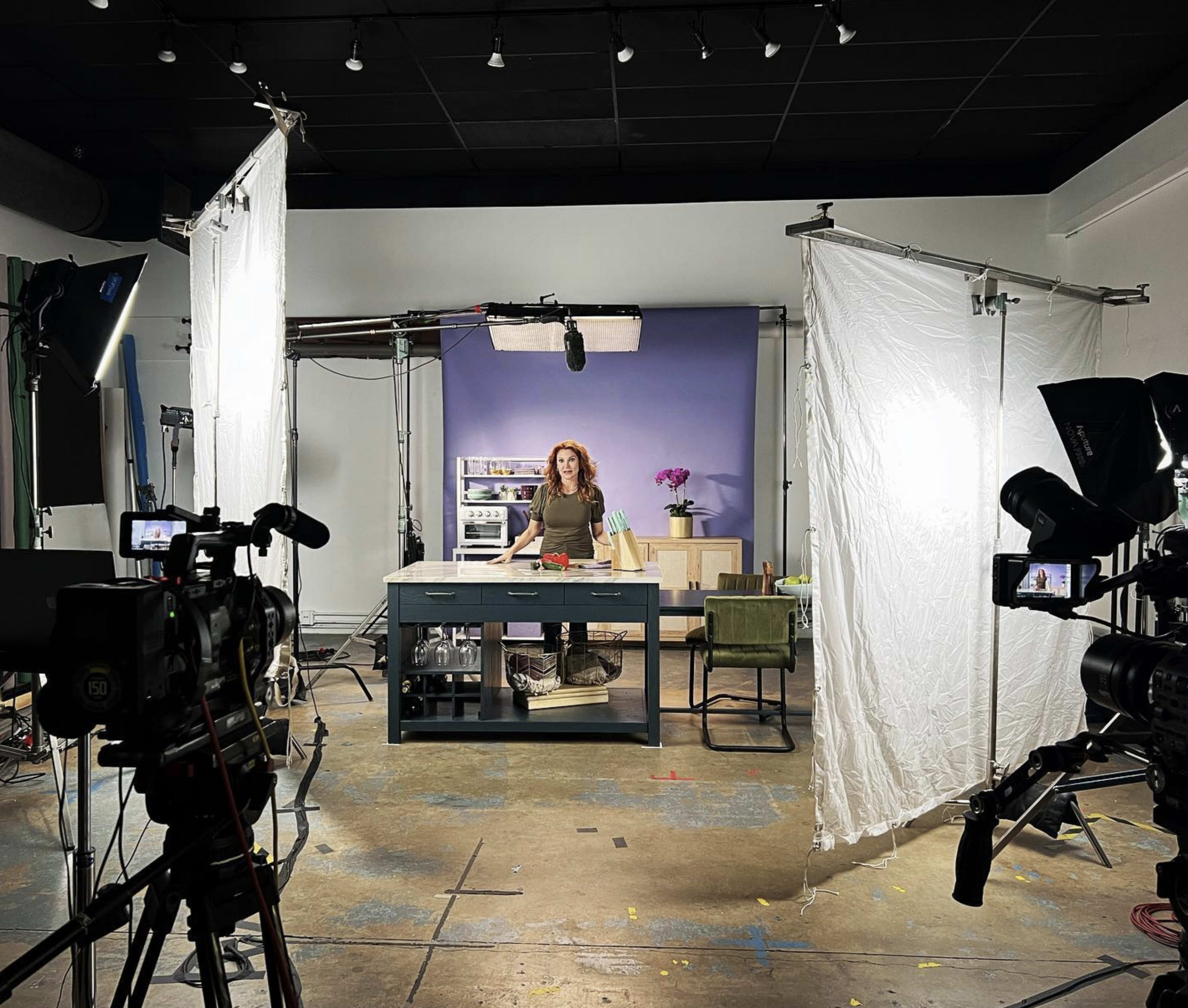 A video production set features a woman standing behind a table surrounded by cameras, lights, and backdrop elements.