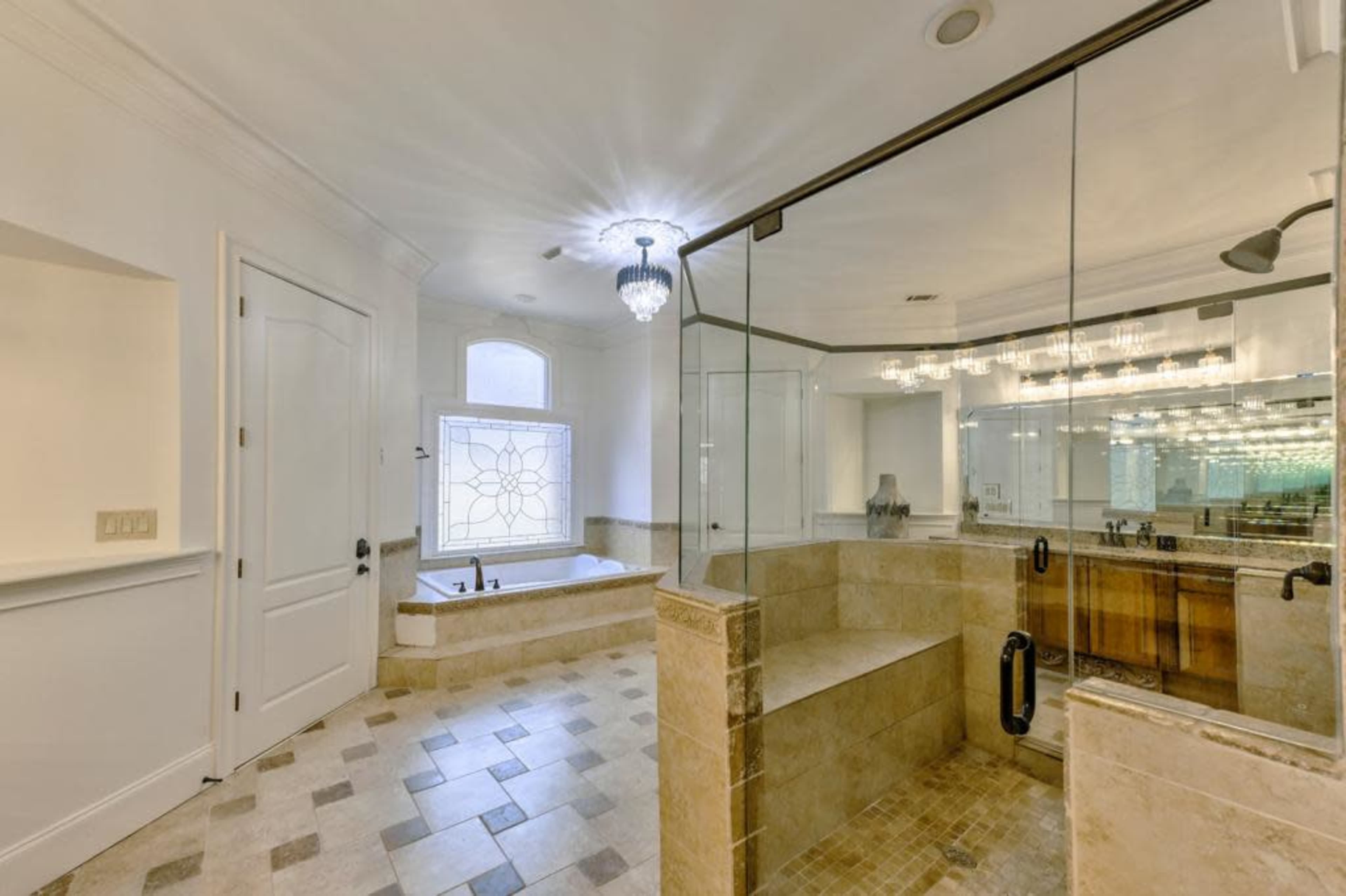 A spacious bathroom featuring a glass-enclosed shower, a large soaking tub by a window, and a double vanity with multiple light fixtures.