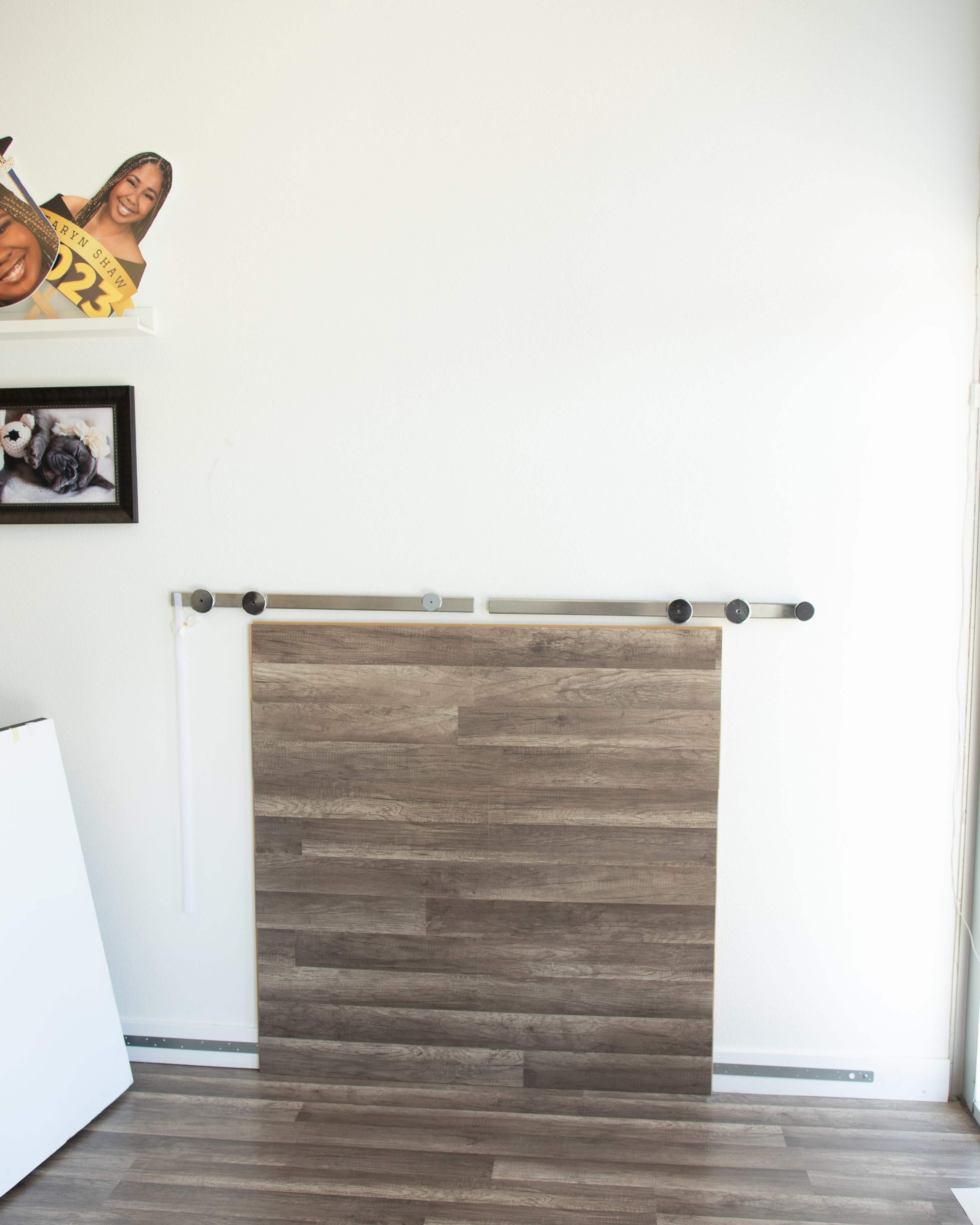 The image shows a wall with a wooden panel mounted on a track, alongside a few framed pictures and a cardboard cutout on a shelf.