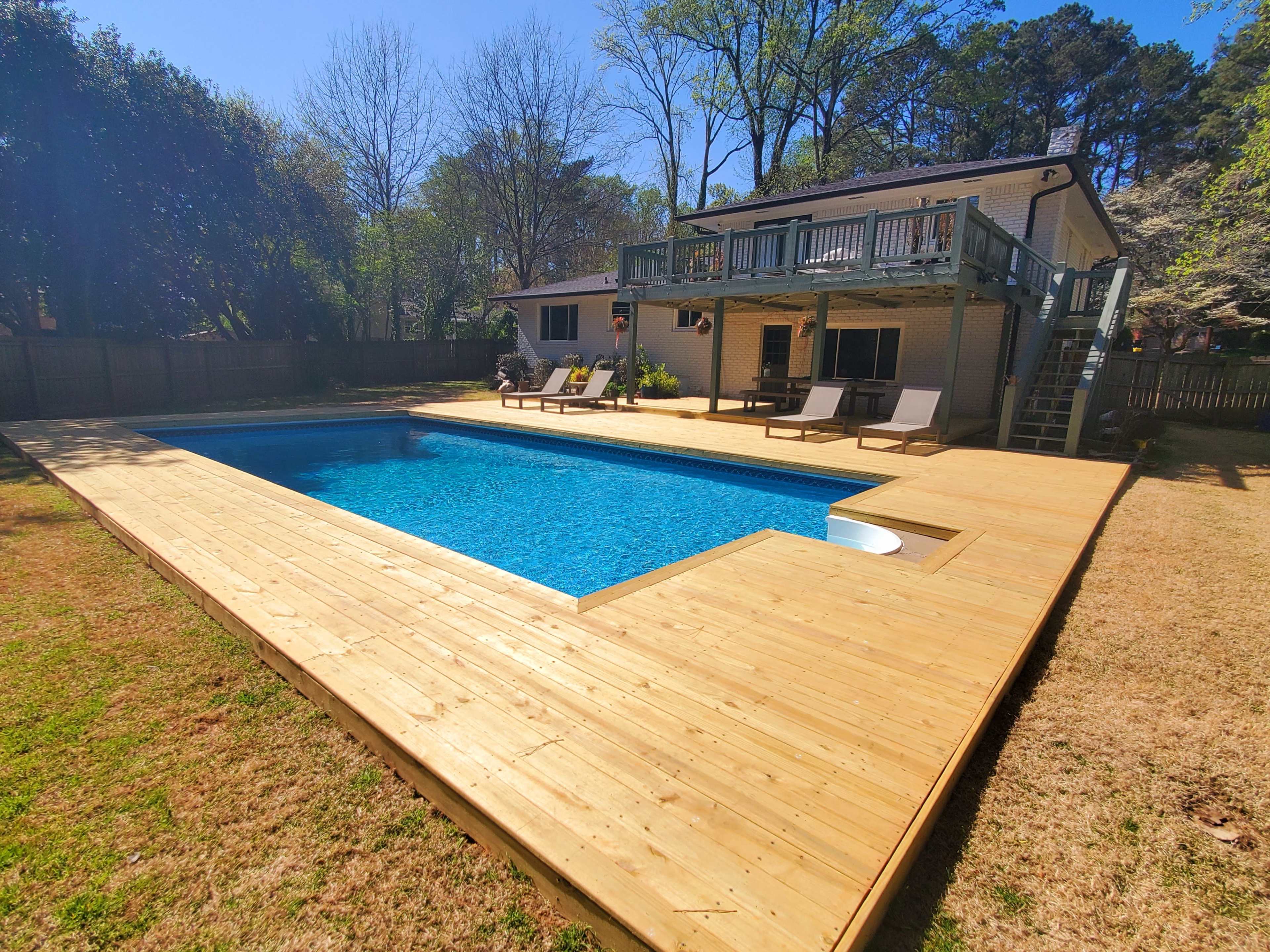 The image shows a backyard swimming pool surrounded by a wooden deck and loungers, with a house in the background and trees nearby.