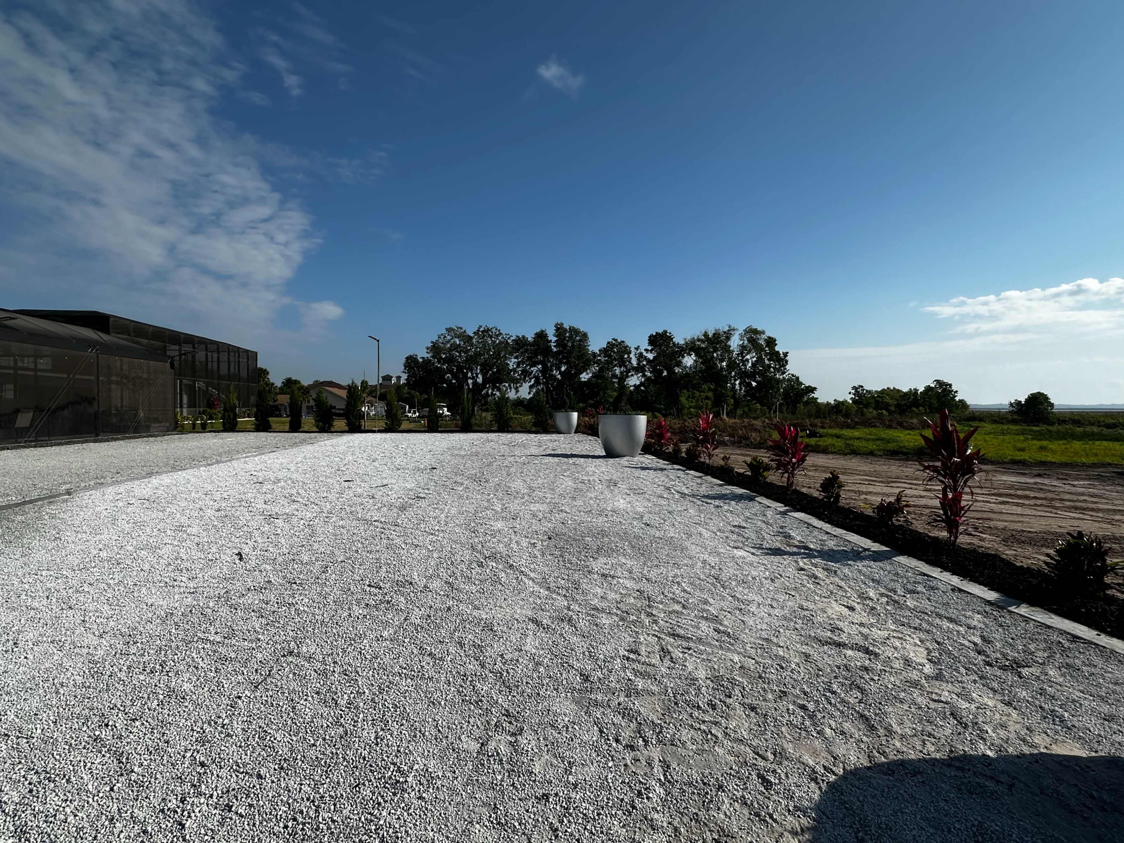 A gravel area is bordered by plants, with a clear sky and green fields in the background.
