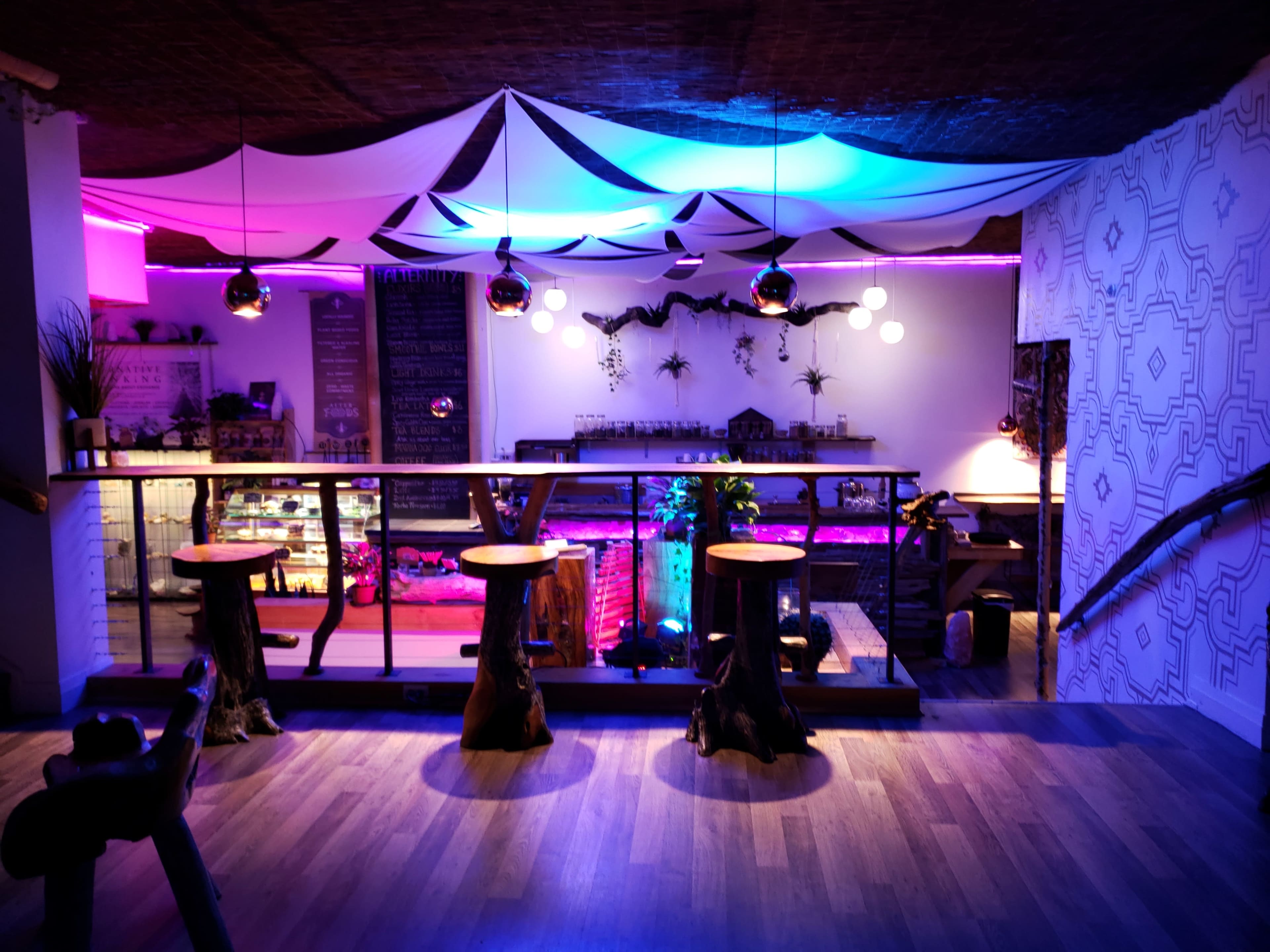 The image shows a modern café interior with a bar area, featuring unique wooden stools, ambient lighting, and decorative plants.