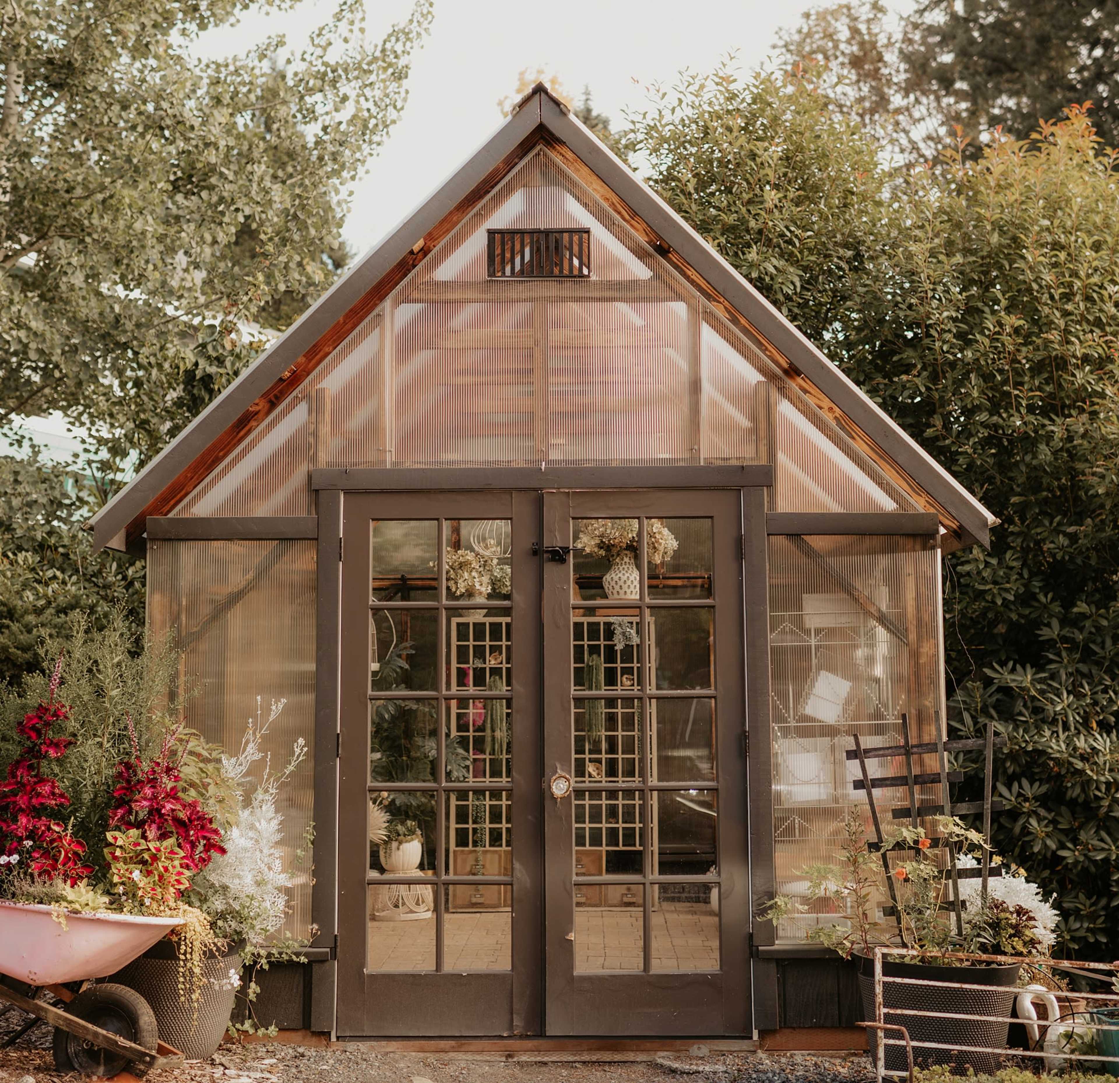 The image shows a greenhouse with a wooden frame and glass panels, featuring double doors and surrounded by various plants and flowers.