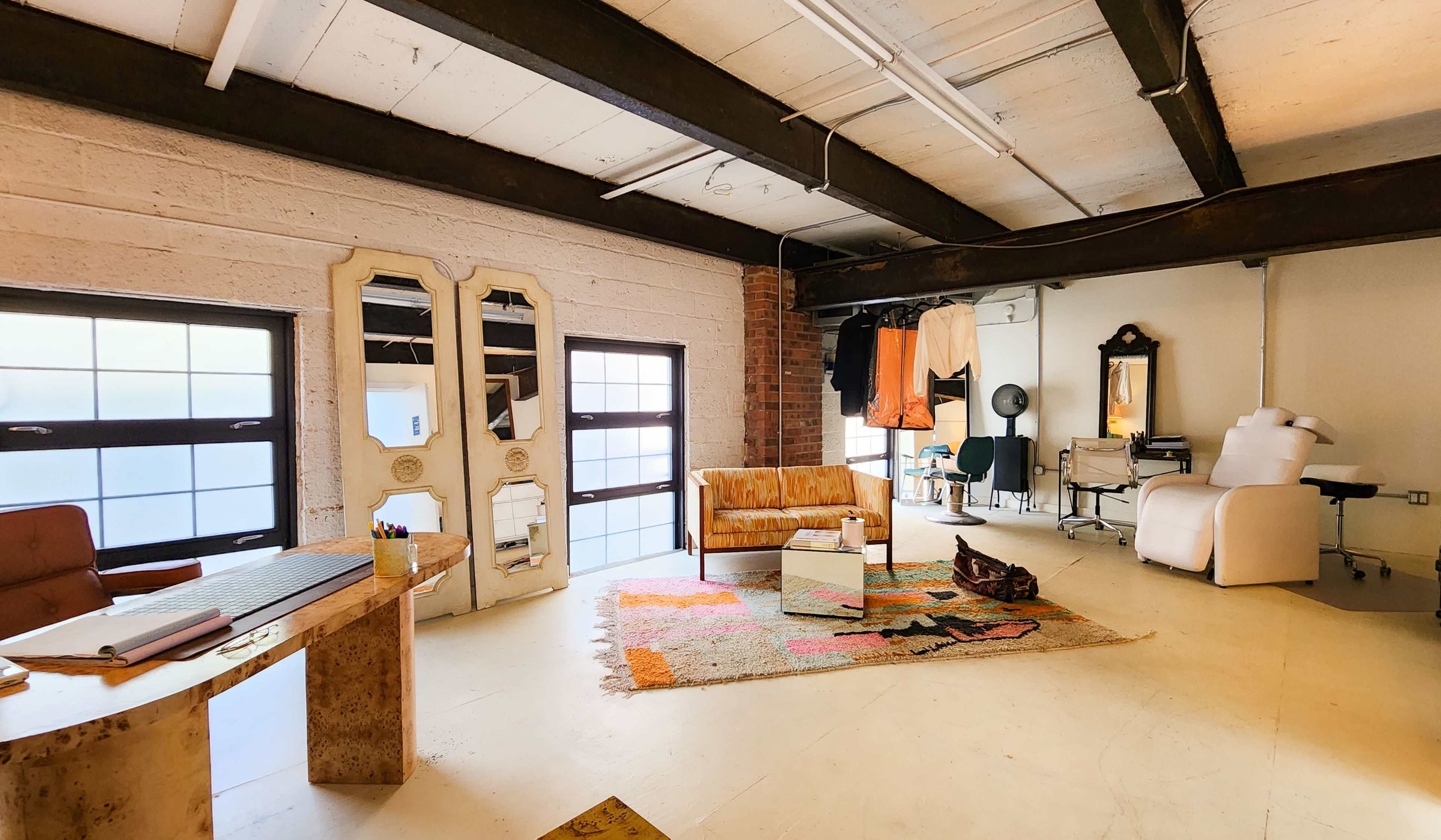 A spacious interior of a room with large windows, featuring a seating area, a desk, various mirrors, and a styling chair.