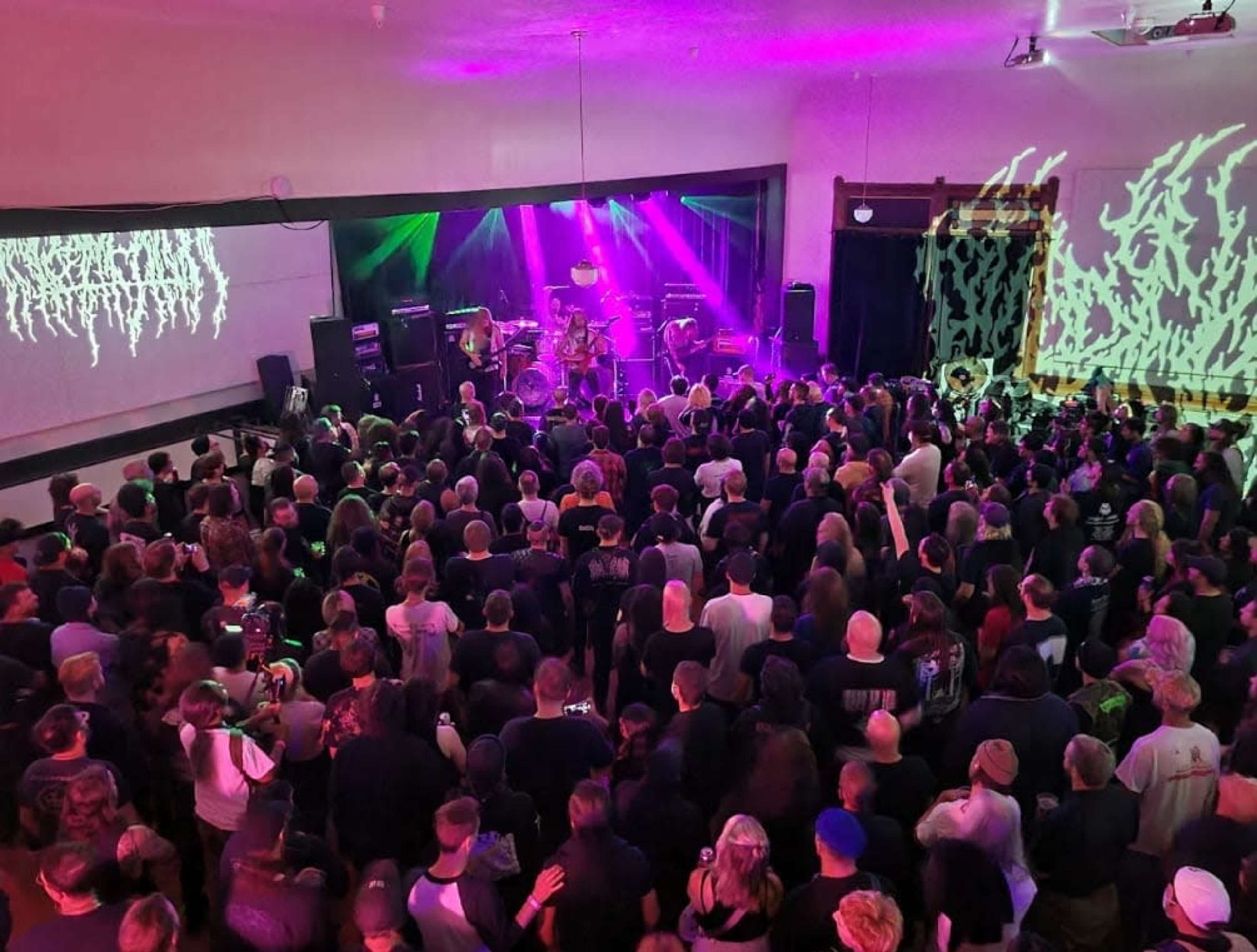 A large crowd watches a band performing on stage with colorful lights and visual projections in a concert venue.