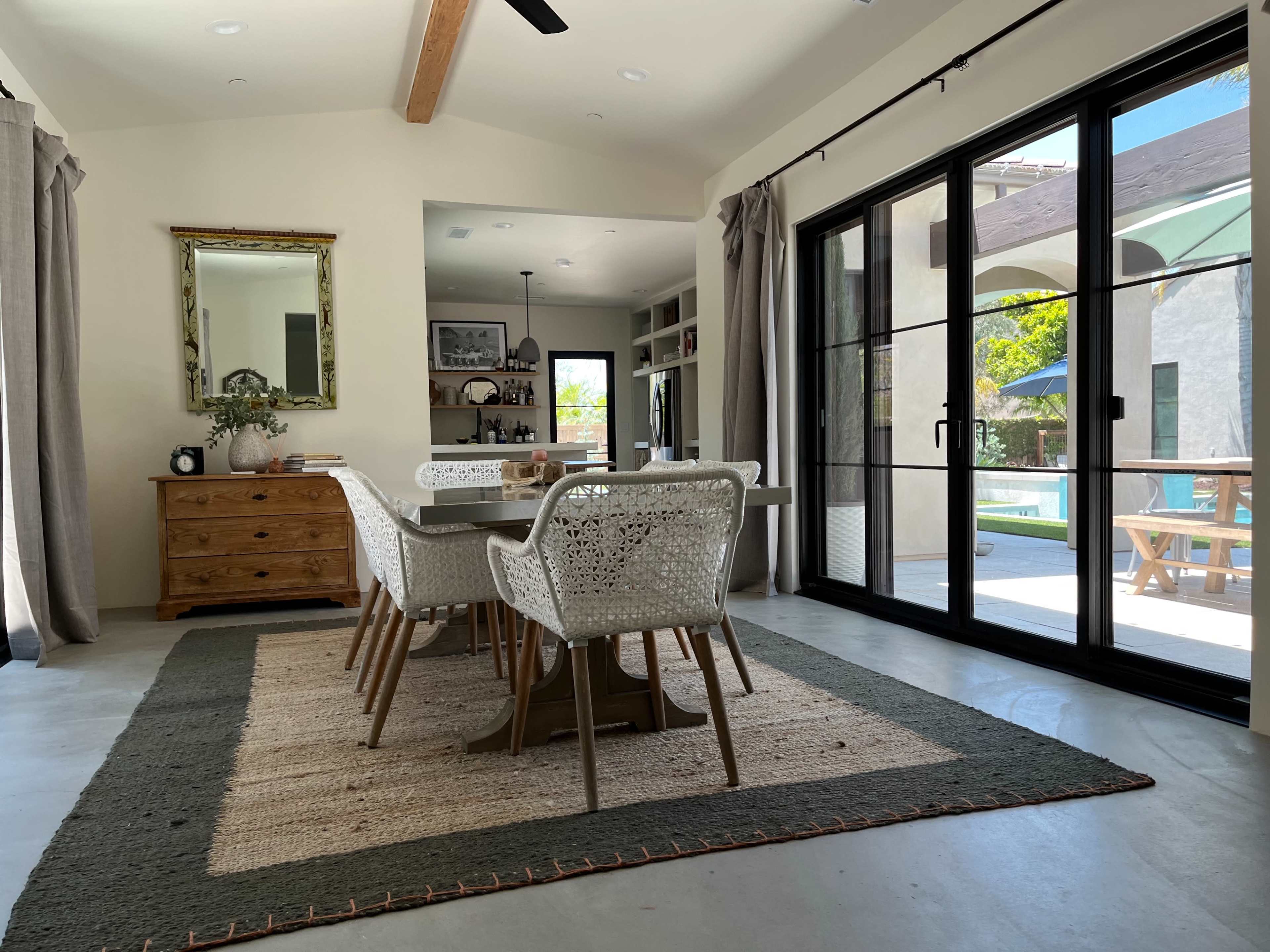 A dining area features a rectangular table surrounded by woven chairs, with a view of a patio through large glass doors.