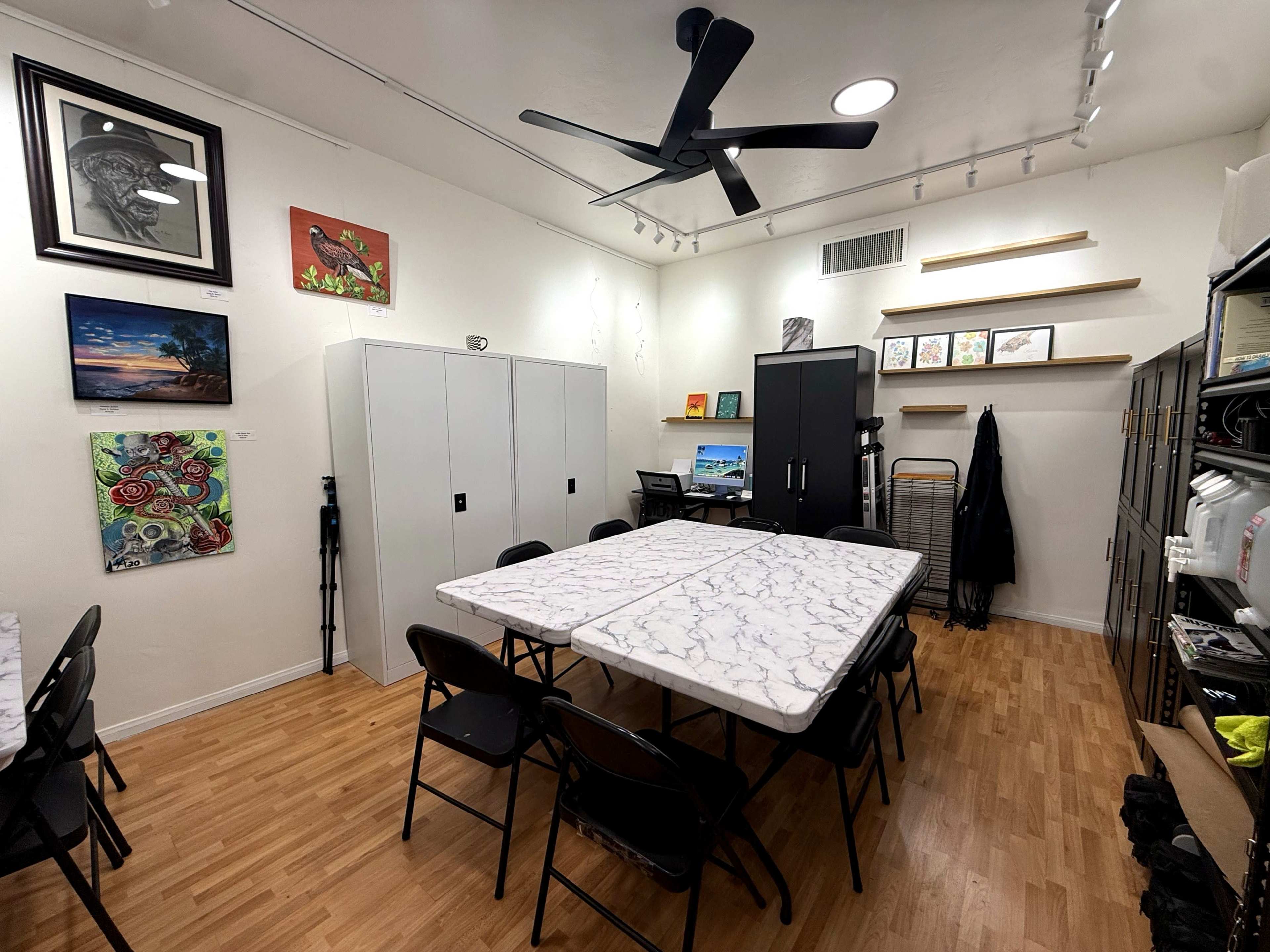 The image shows a well-lit room featuring a long table with chairs, storage cabinets, artwork on the walls, and a desk in the corner.