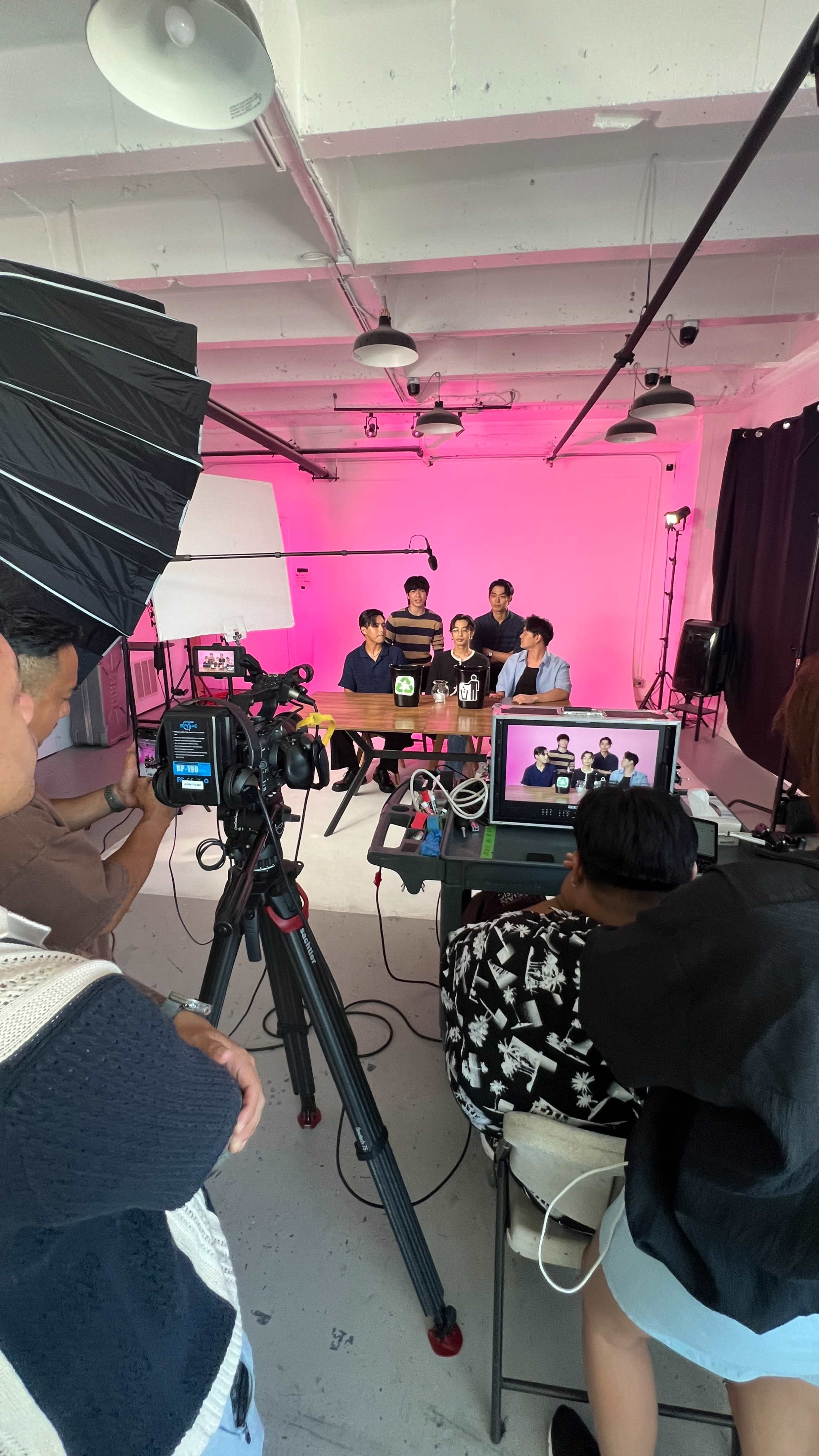 A group of five individuals is seated at a table in a brightly lit studio with pink walls, while a camera and crew set up for filming.