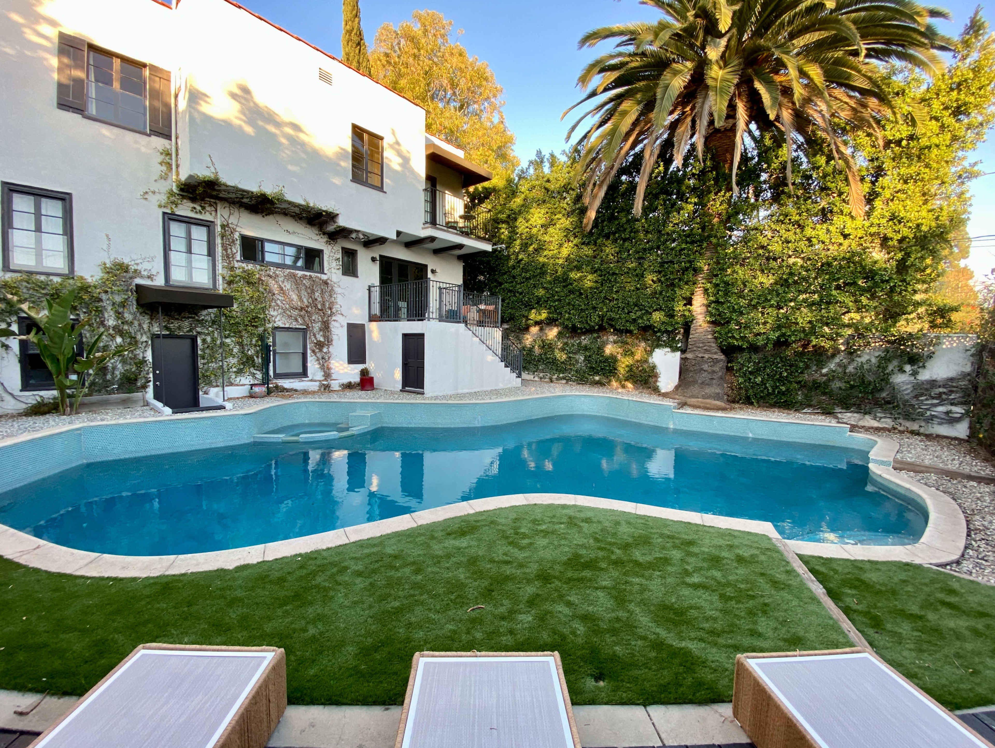 A sparkling blue swimming pool surrounded by a landscaped area featuring palm trees and a multi-level house in the background.