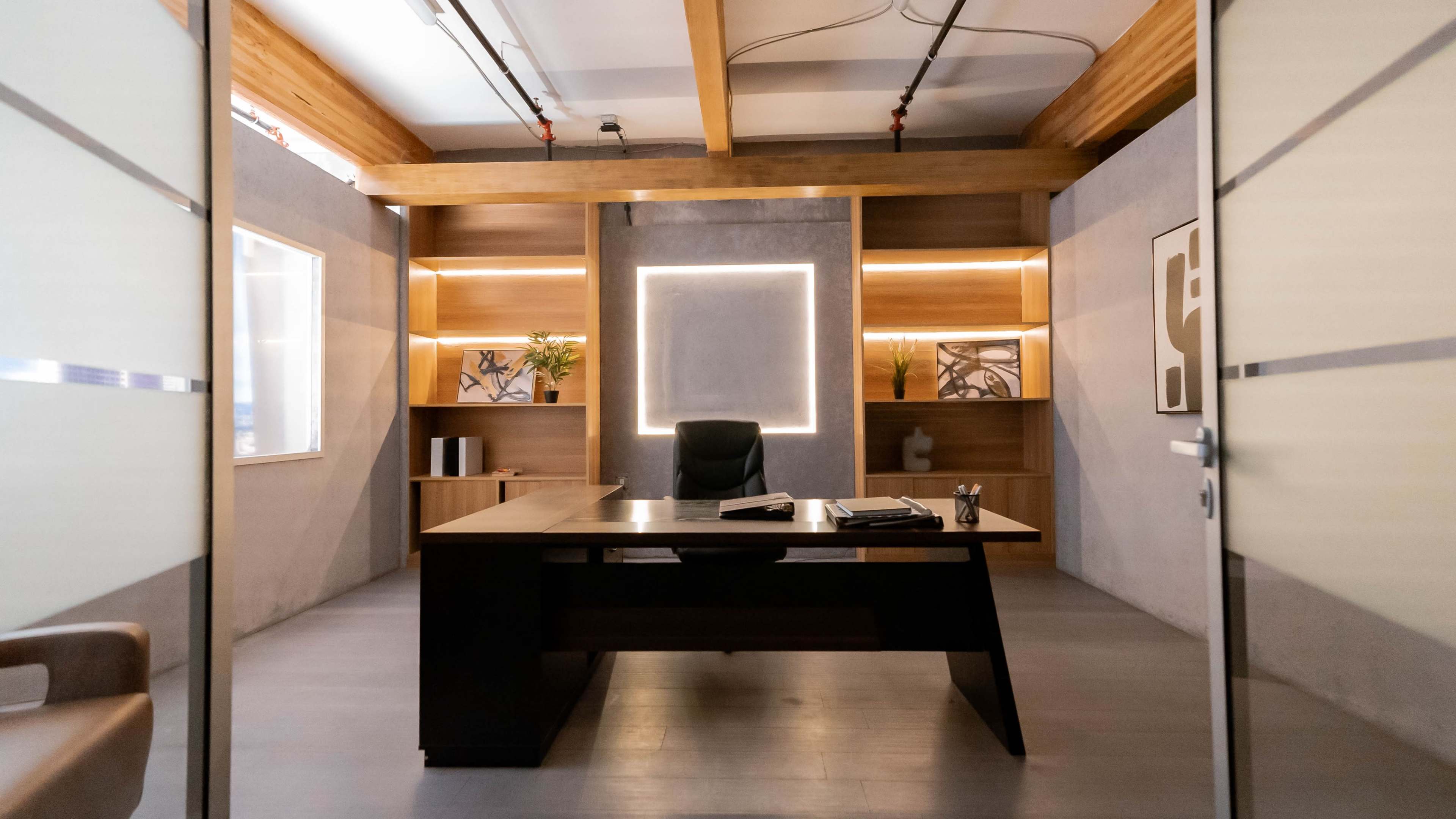 The image shows a modern office space with a central desk, shelving with decorative items, and large glass doors.