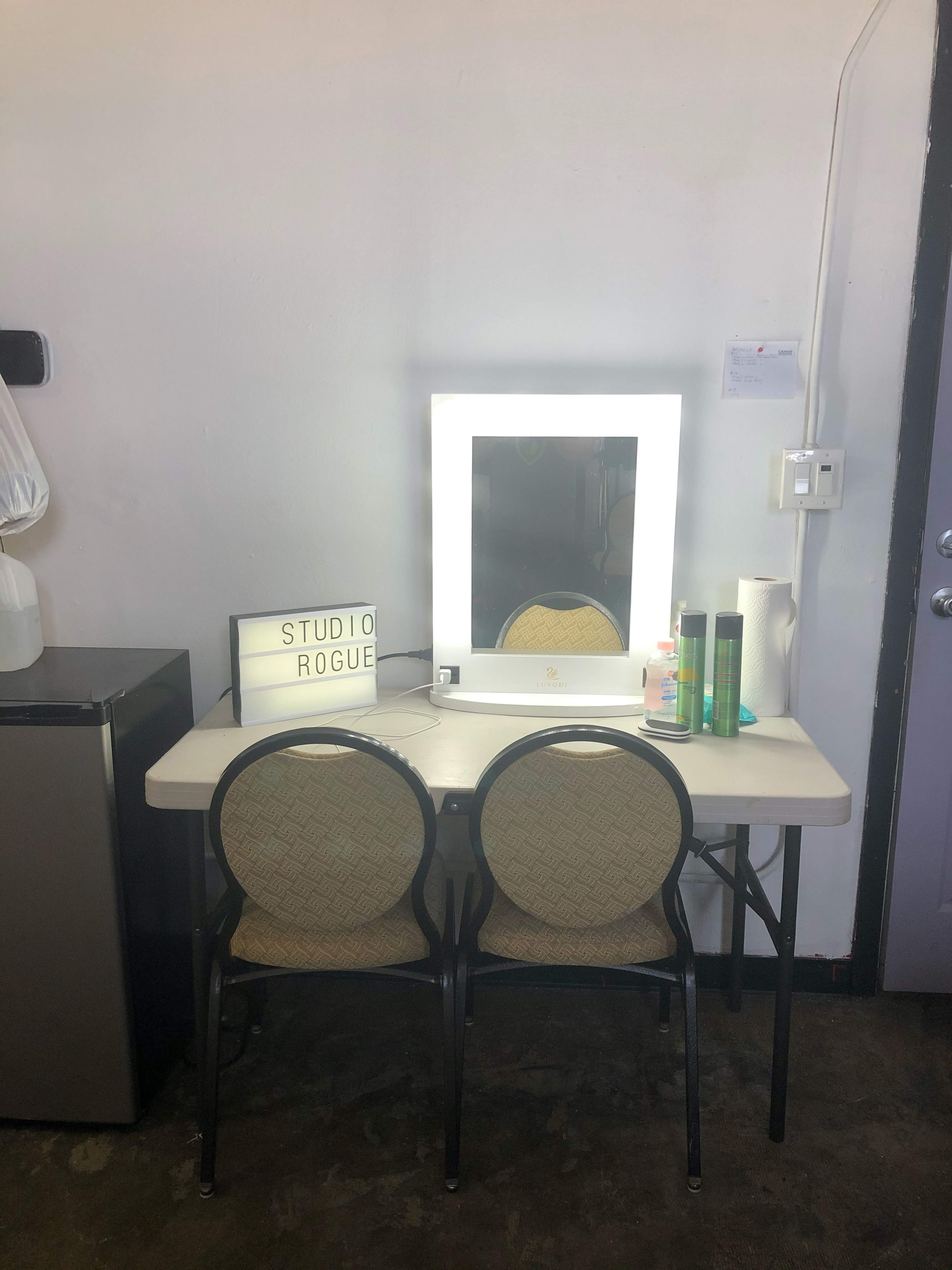 A clean, simple vanity setup with a lighted mirror, two chairs, and various beauty products on a table against a plain wall.