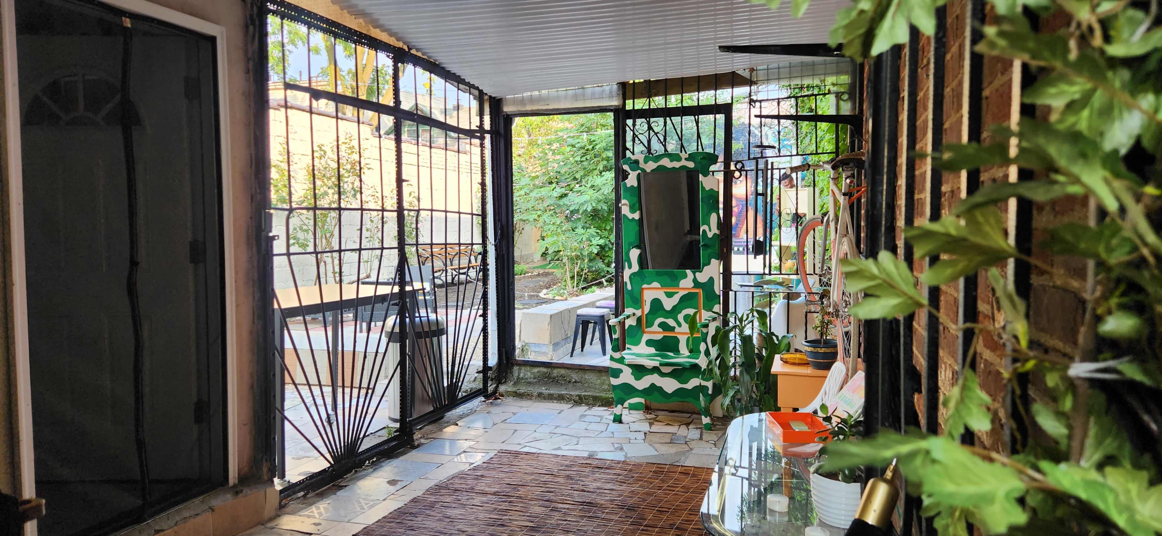 The image shows a narrow outdoor corridor with metal gates leading into a garden area, surrounded by plants and various pieces of outdoor furniture.