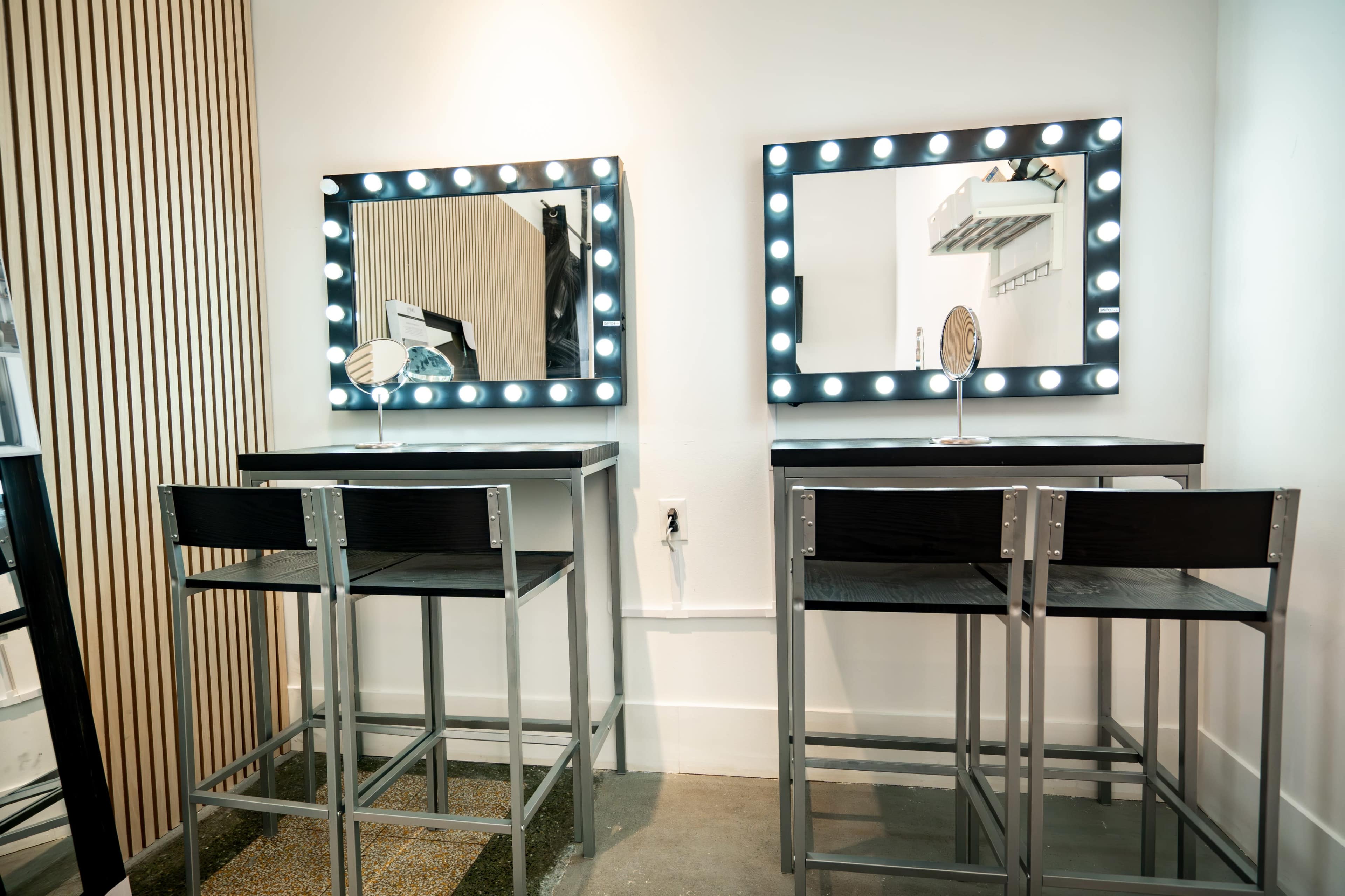 The image features two illuminated vanity mirrors mounted on a wall, with matching black stools positioned in front of each mirror.