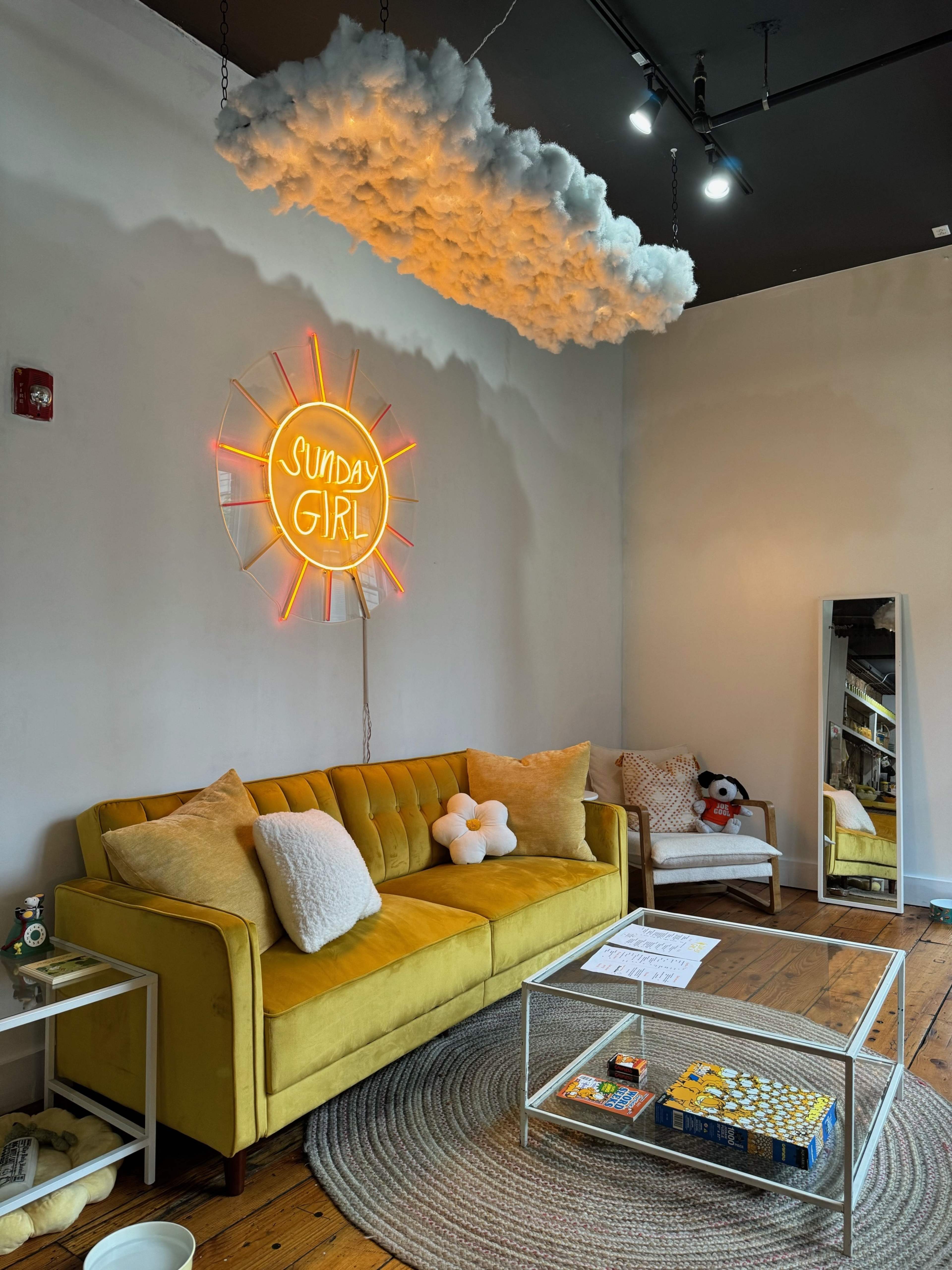 A bright yellow sofa is positioned under a neon sign that reads "SUNDAY GIRL," with a cloud-shaped decoration above it in a stylish, minimalist room.