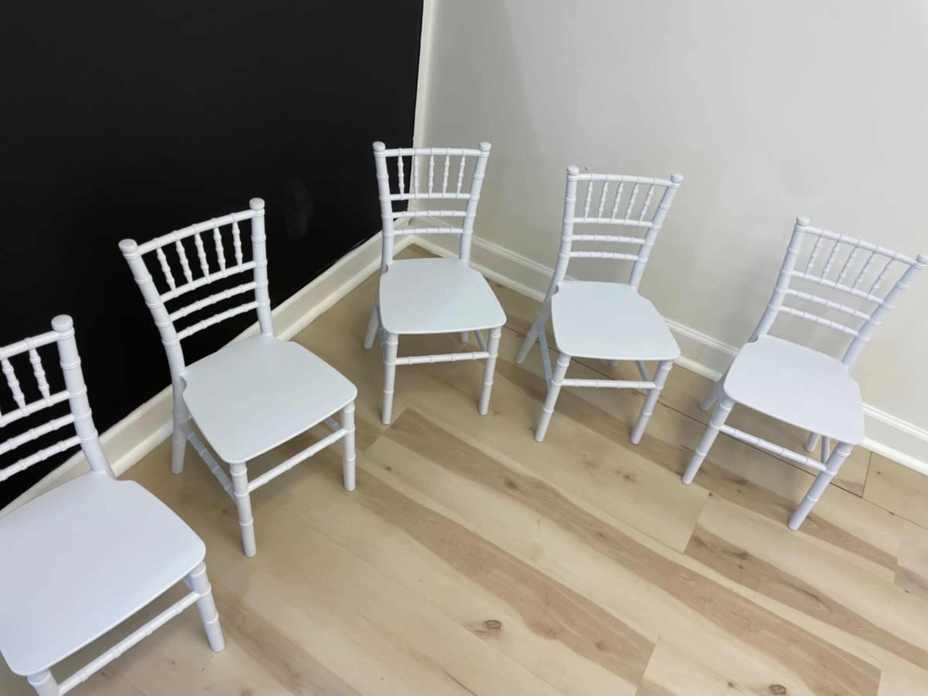 Five white chairs are arranged in a row on a wooden floor against a black wall.